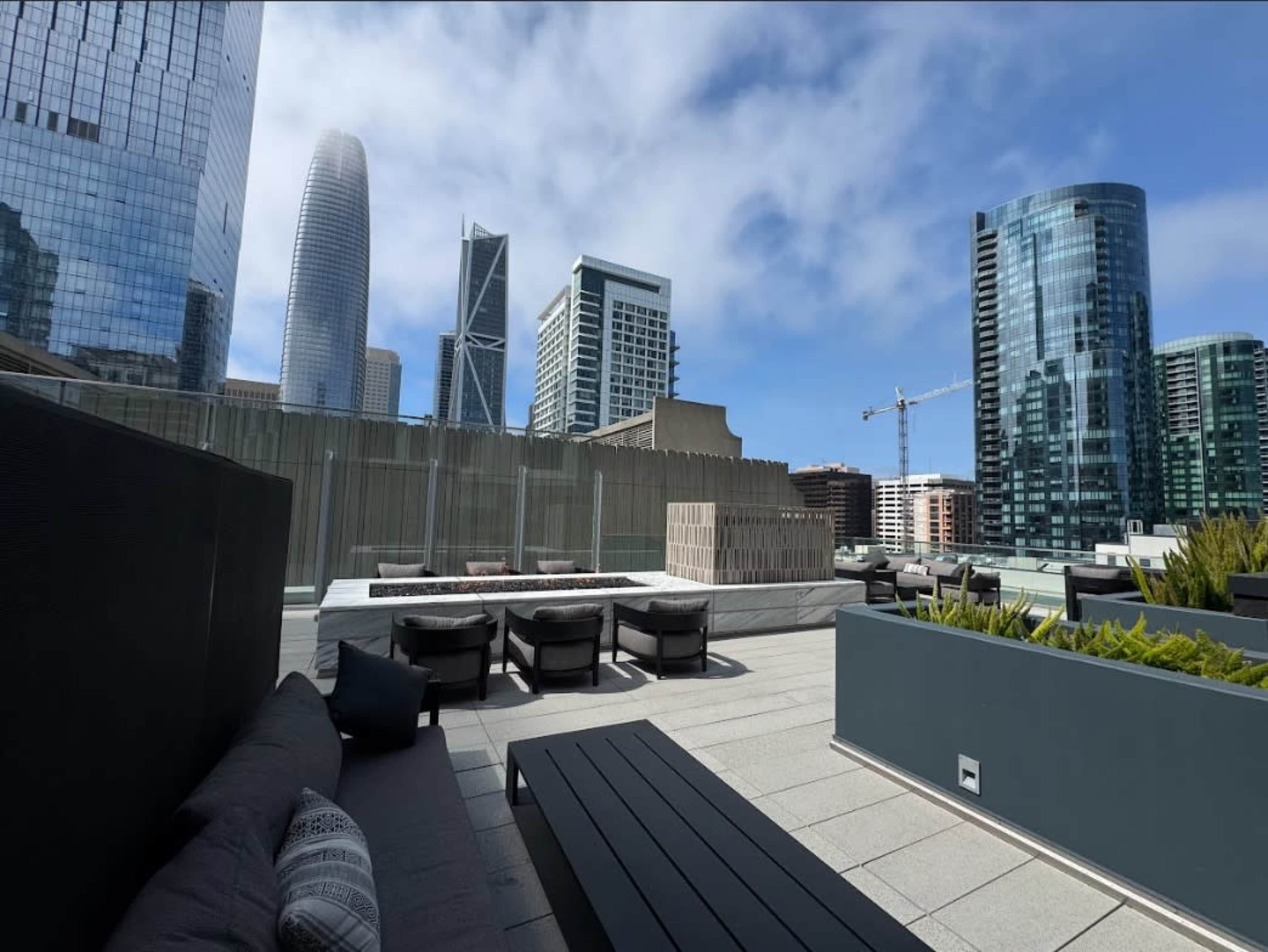 A modern rooftop terrace features sleek seating and planters, surrounded by tall buildings under a partly cloudy sky.