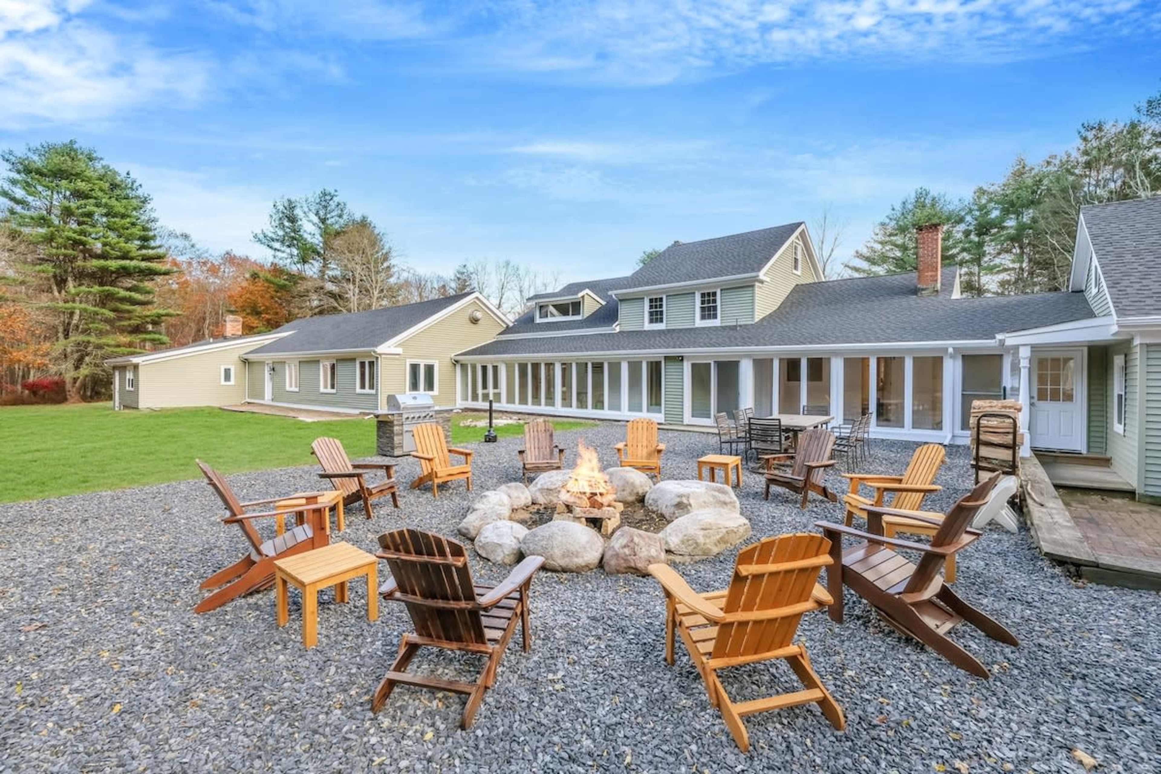 A circular arrangement of wooden chairs surrounds a fire pit in a gravel yard, with a spacious house and trees in the background.