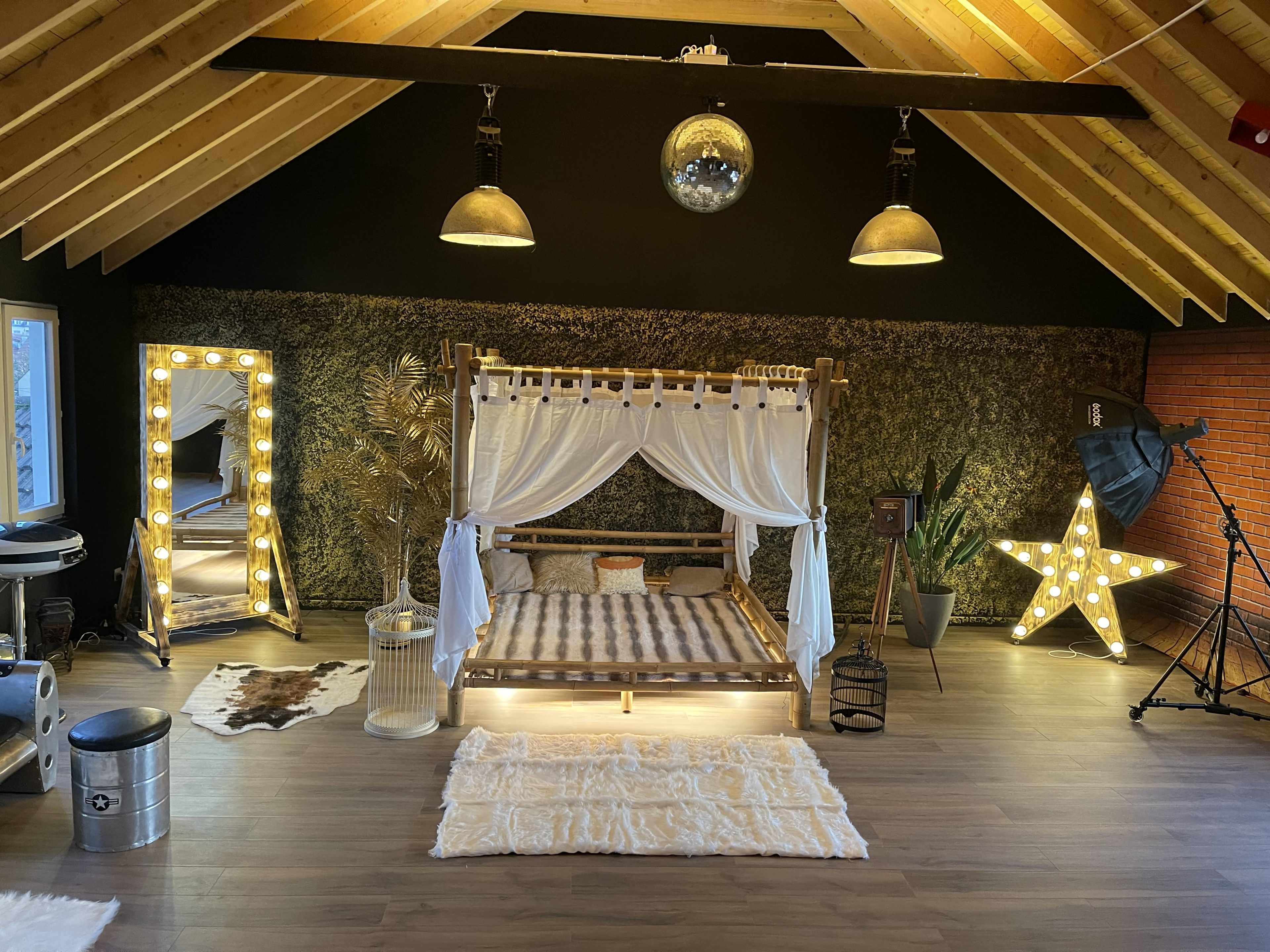The image shows a spacious, stylish bedroom featuring a four-poster bed with white drapes, a large illuminated mirror, and decorative lighting elements against a wooden ceiling.