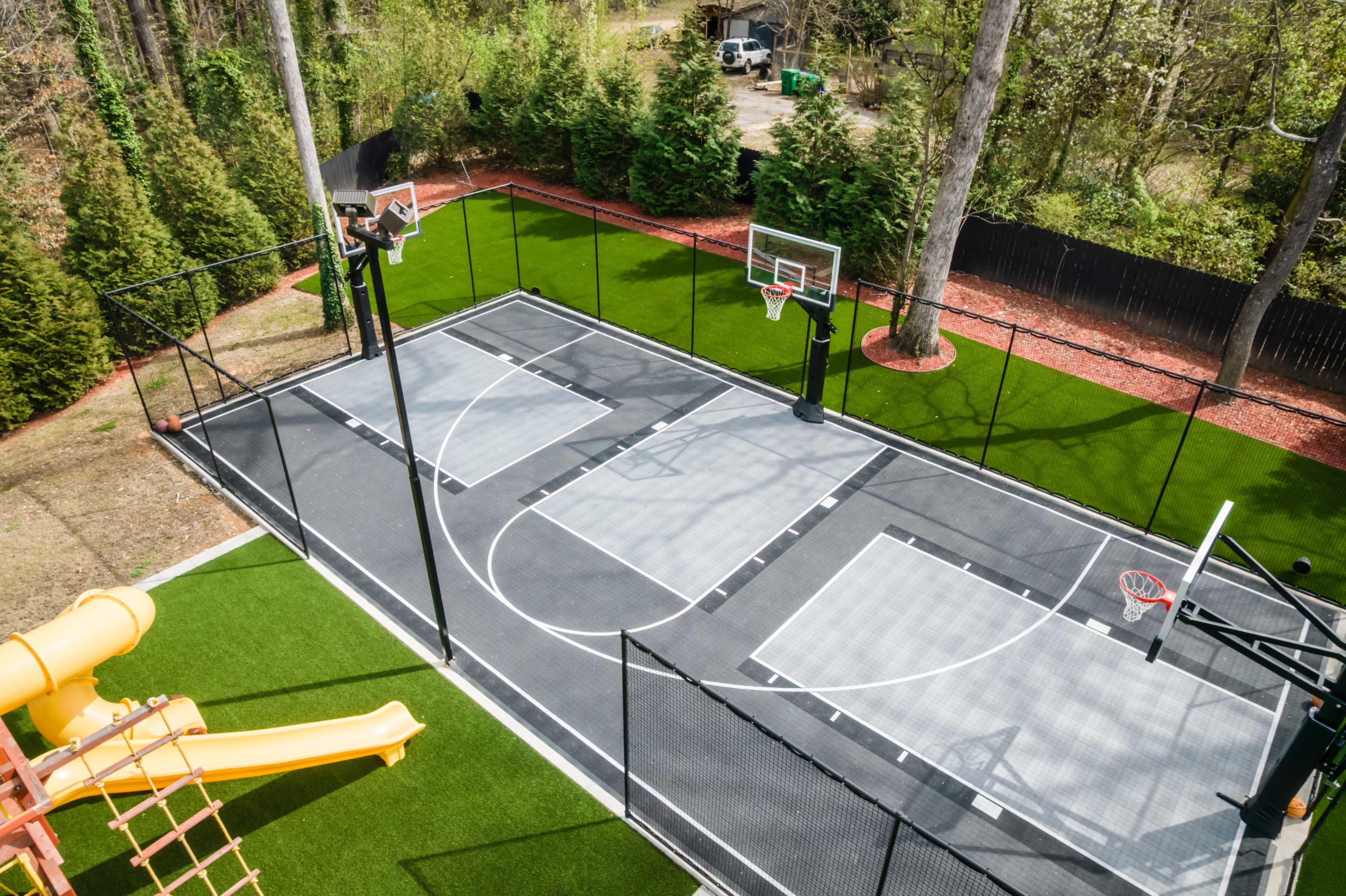 The image shows a fenced outdoor basketball court featuring two hoops, surrounded by green grass and trees, with a yellow playground slide nearby.