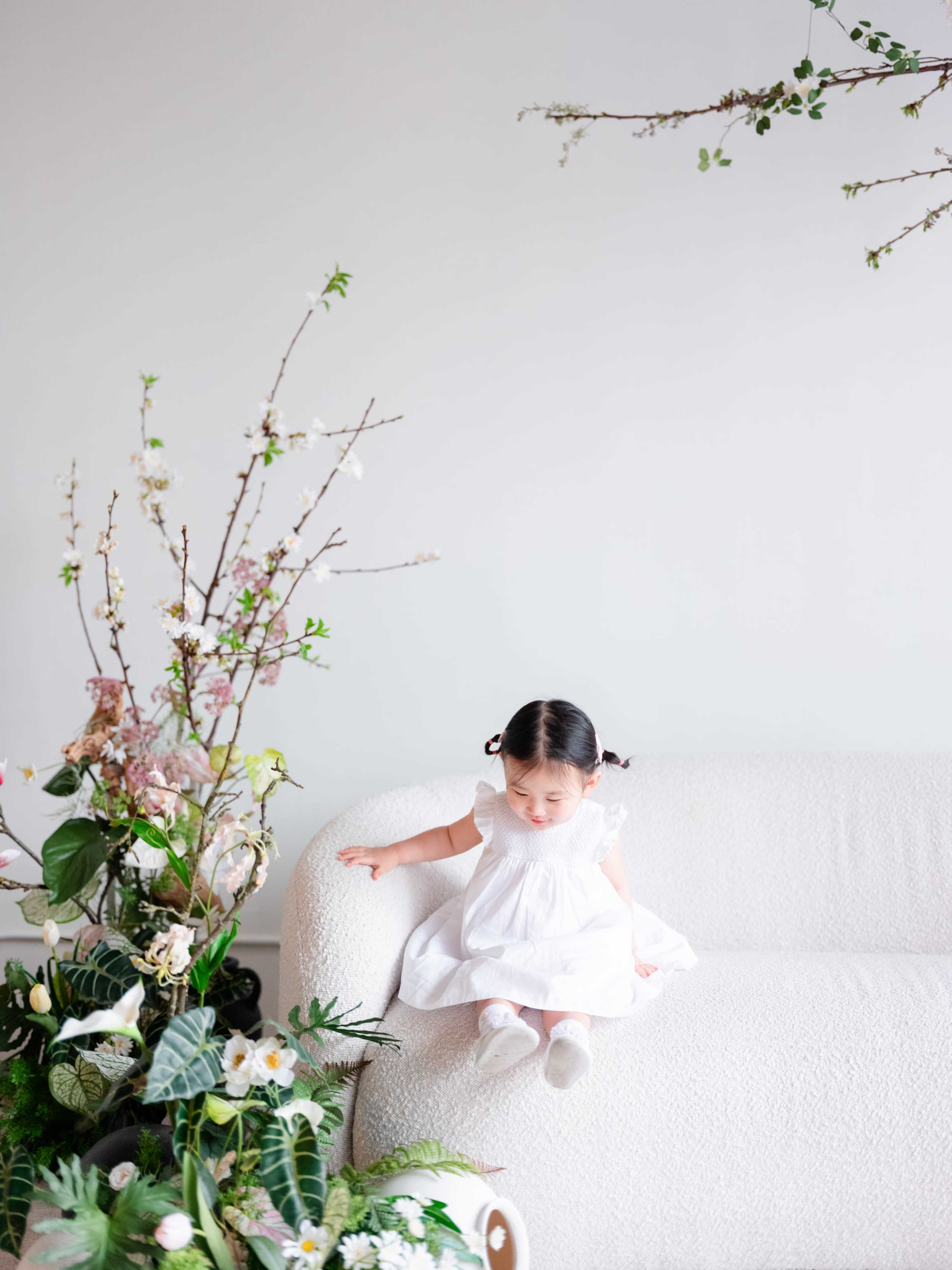 A young child in a white dress sits on a light-colored sofa next to a large floral arrangement.