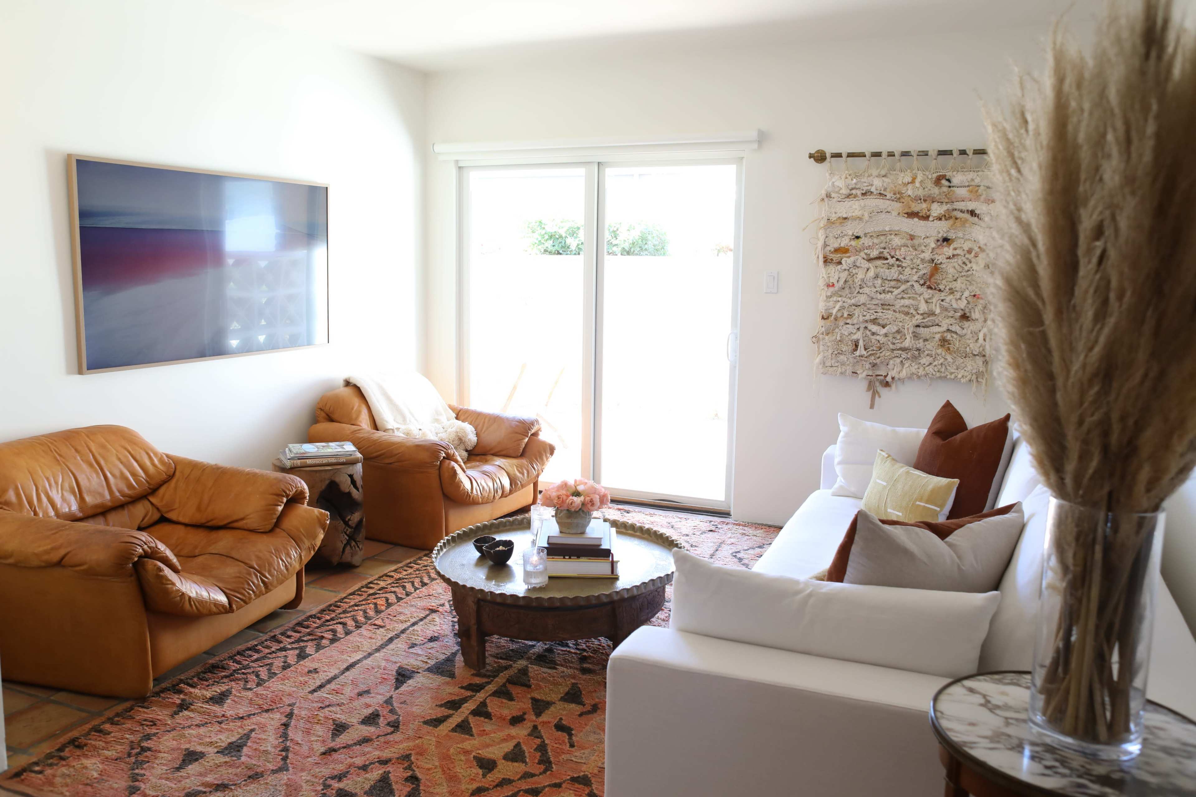 The living room features two brown leather chairs, a white sofa, a round coffee table, and a tapestry hung on the wall, with large windows letting in natural light.
