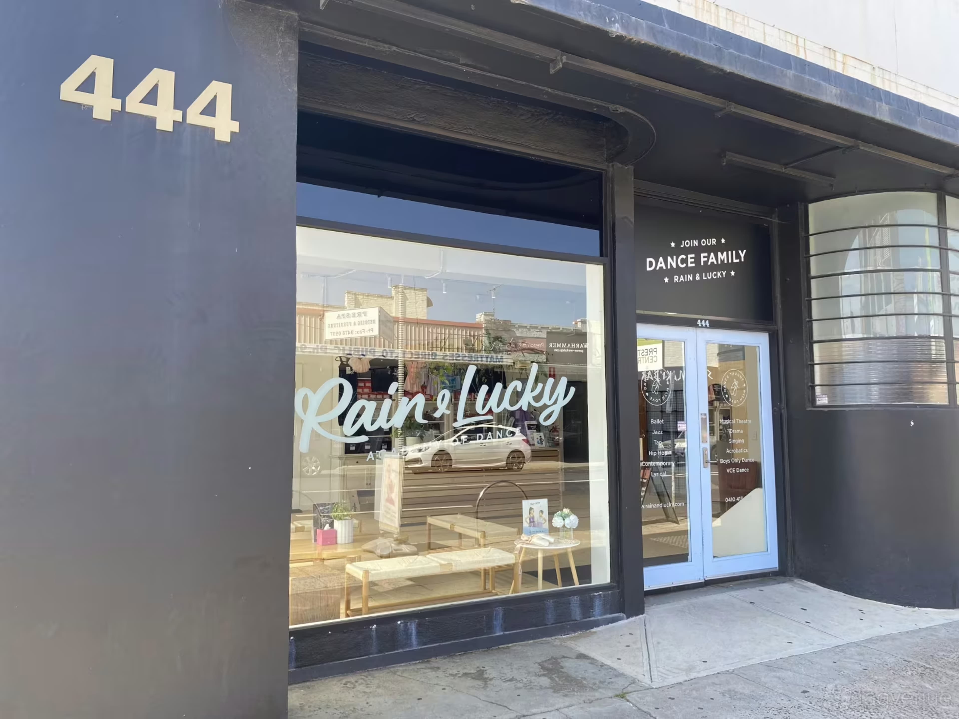 The image shows the storefront of a business named "Rain + Lucky" located at 444, featuring a large window display and a door with signage promoting dance classes.