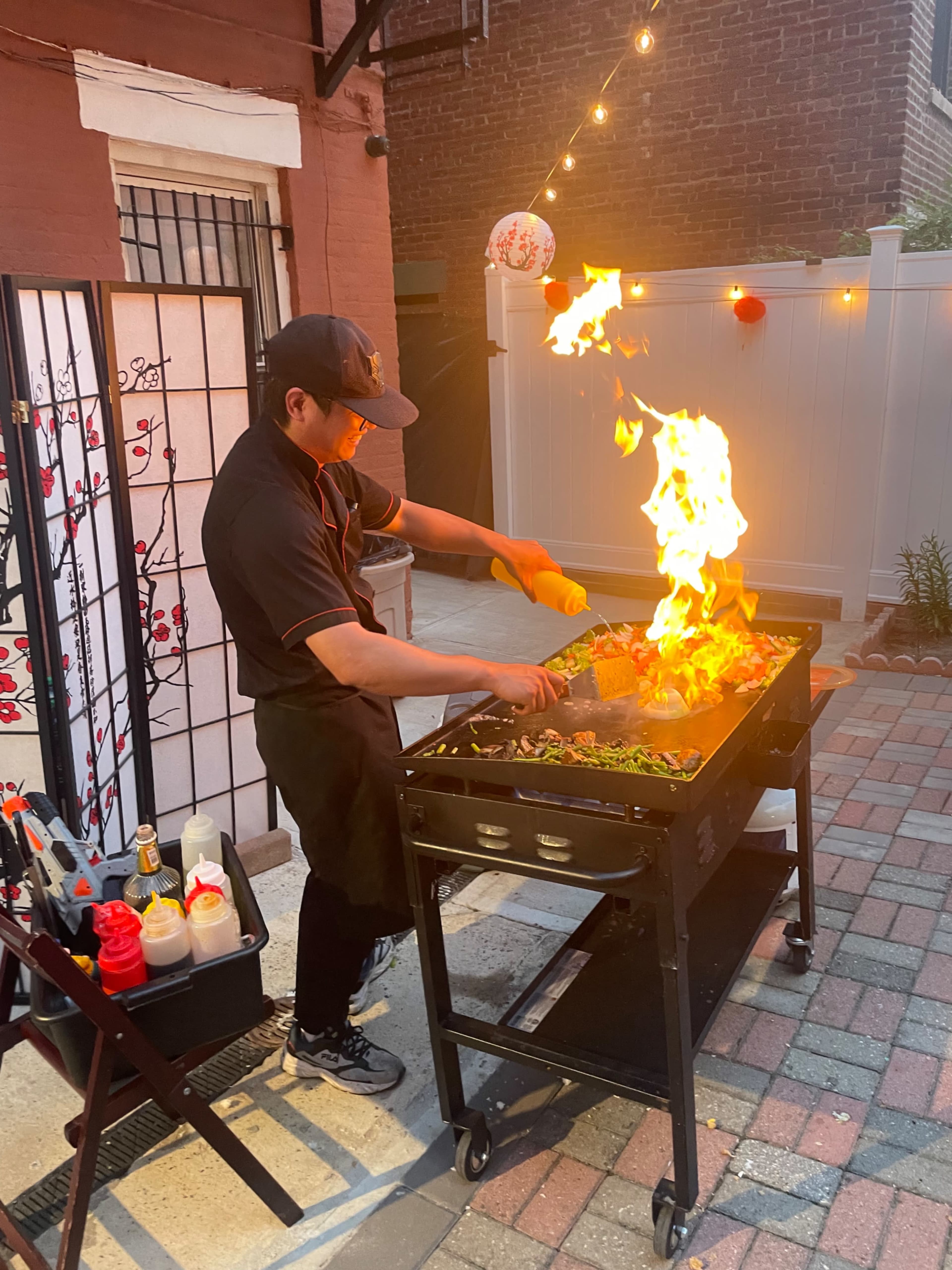 A chef grills food on an outdoor barbecue, with flames rising dramatically as he seasons the meat.