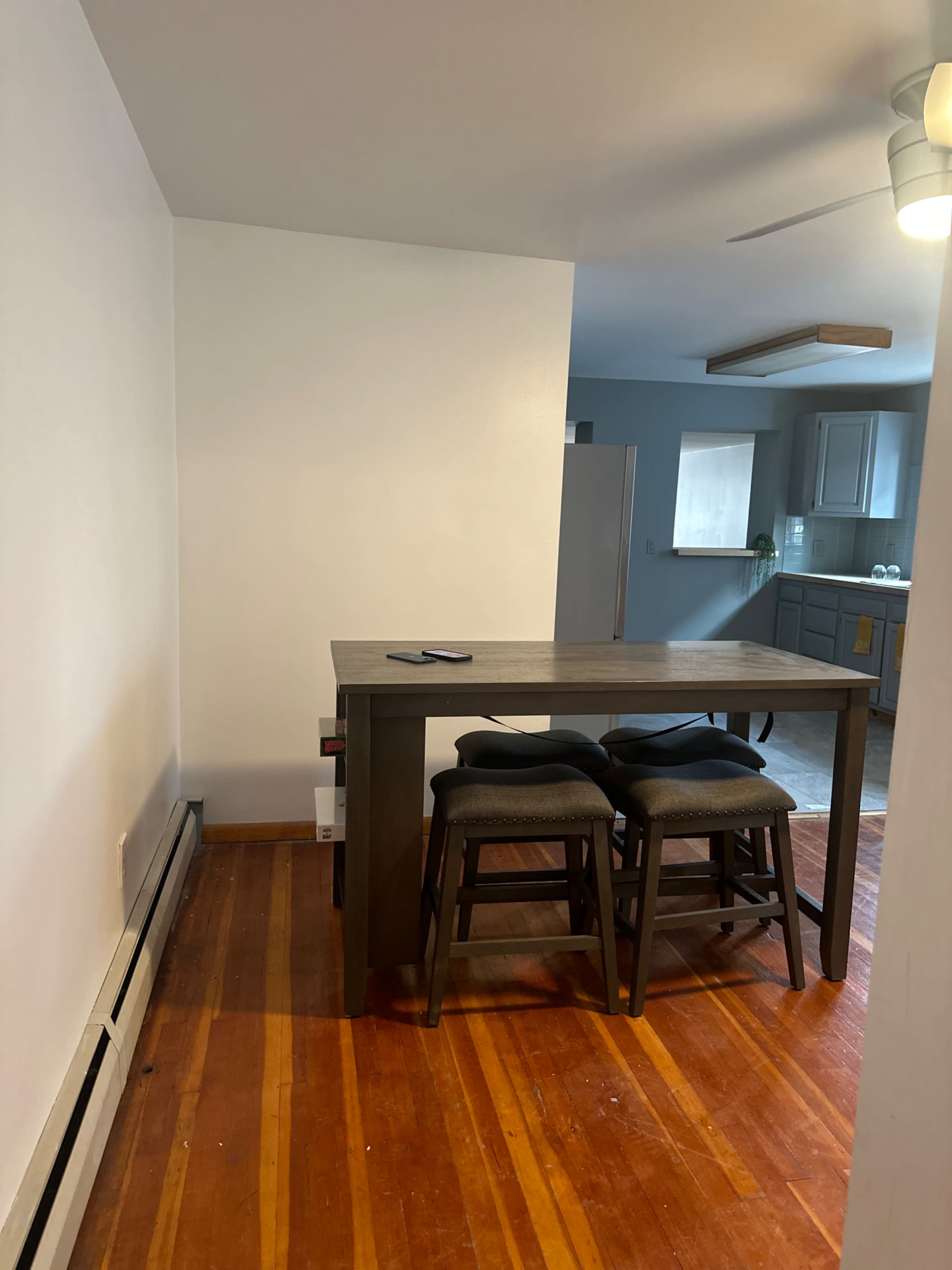 A wooden dining table with two chairs is positioned against a plain wall in a brightly lit room that leads to a kitchen area.
