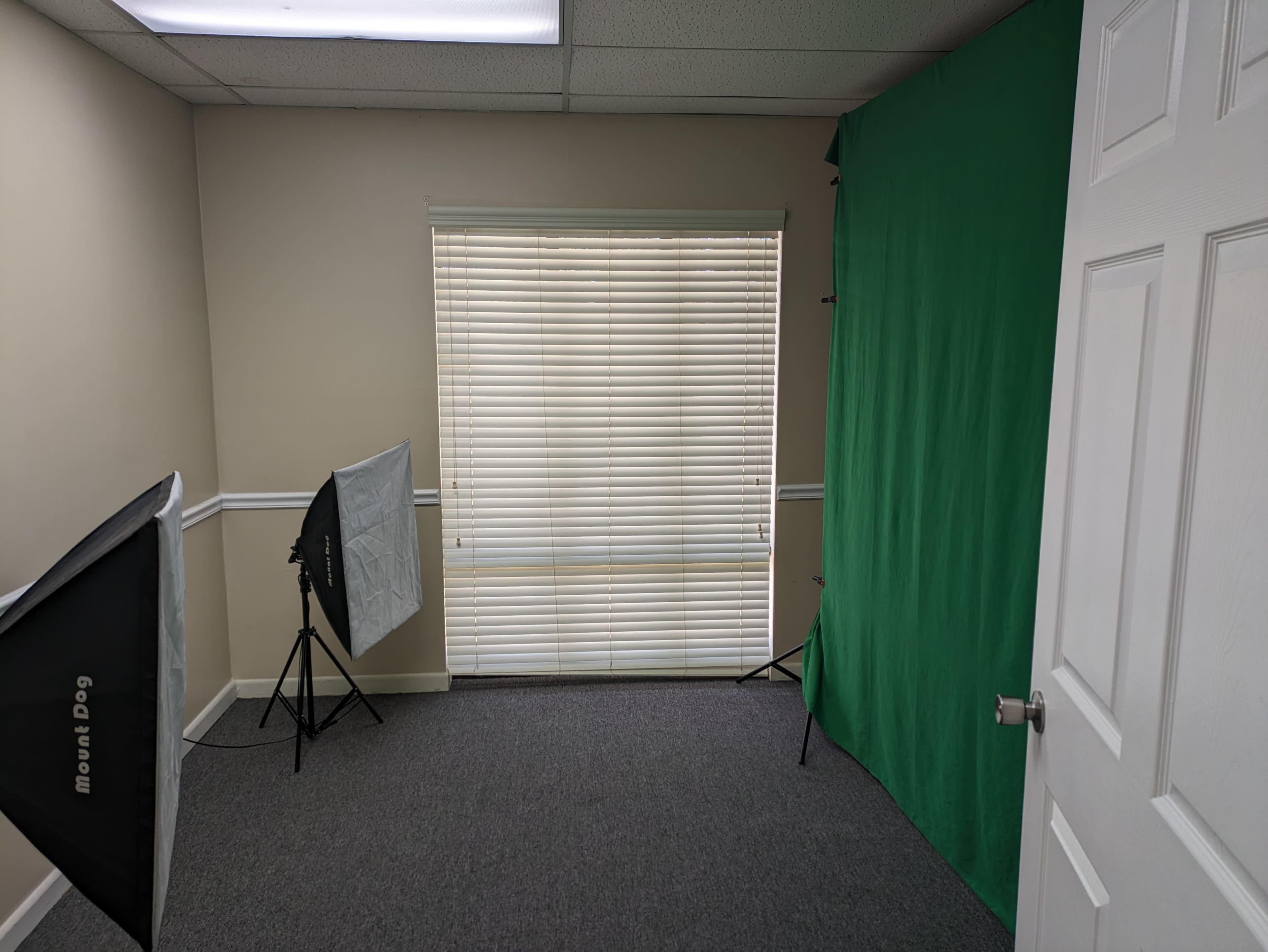 A small room with a green backdrop, a window with blinds, and two softbox lights set up on stands.