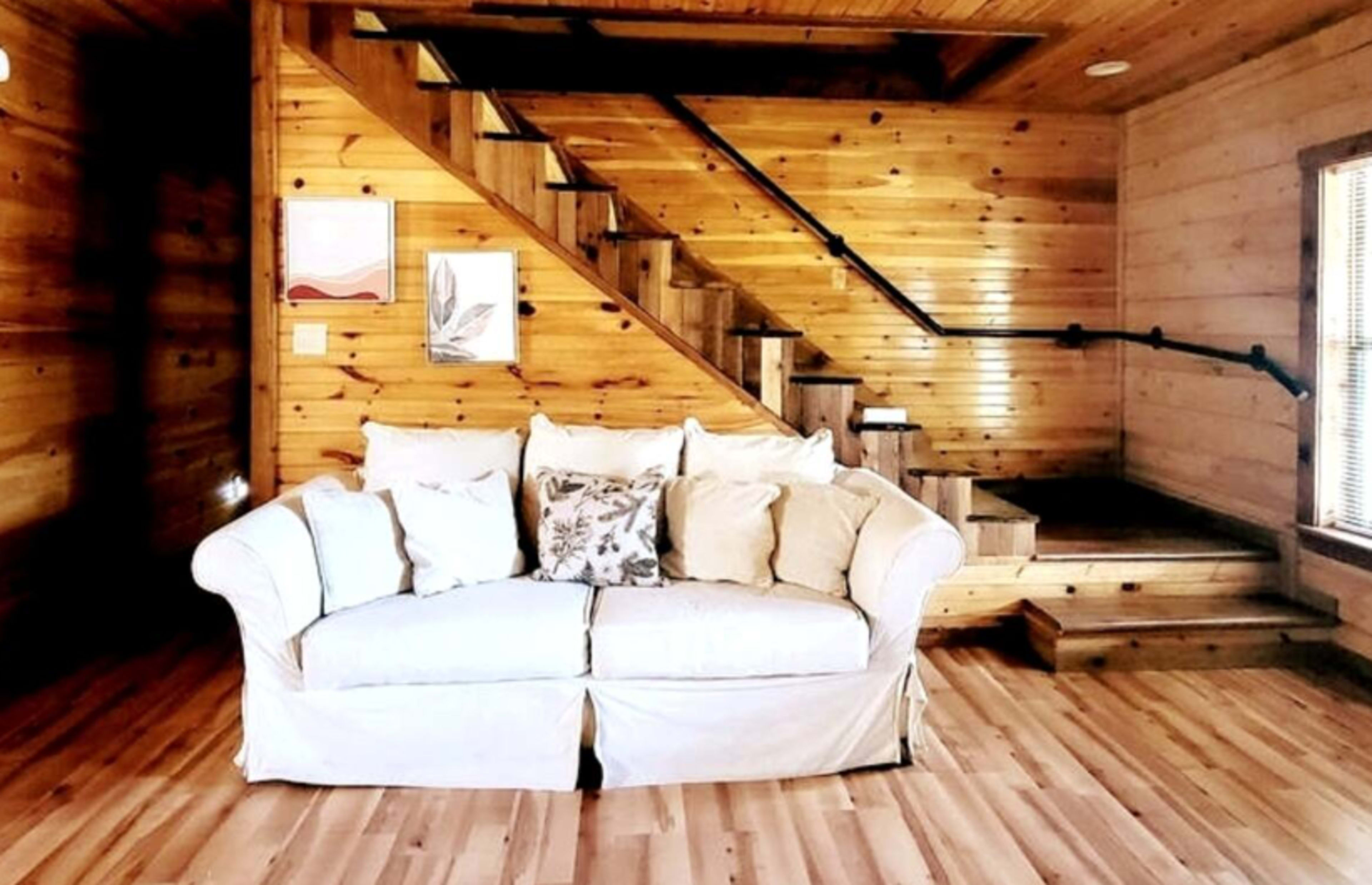 A white sofa with pillows is positioned in a wooden room that features a staircase and natural wood paneling.