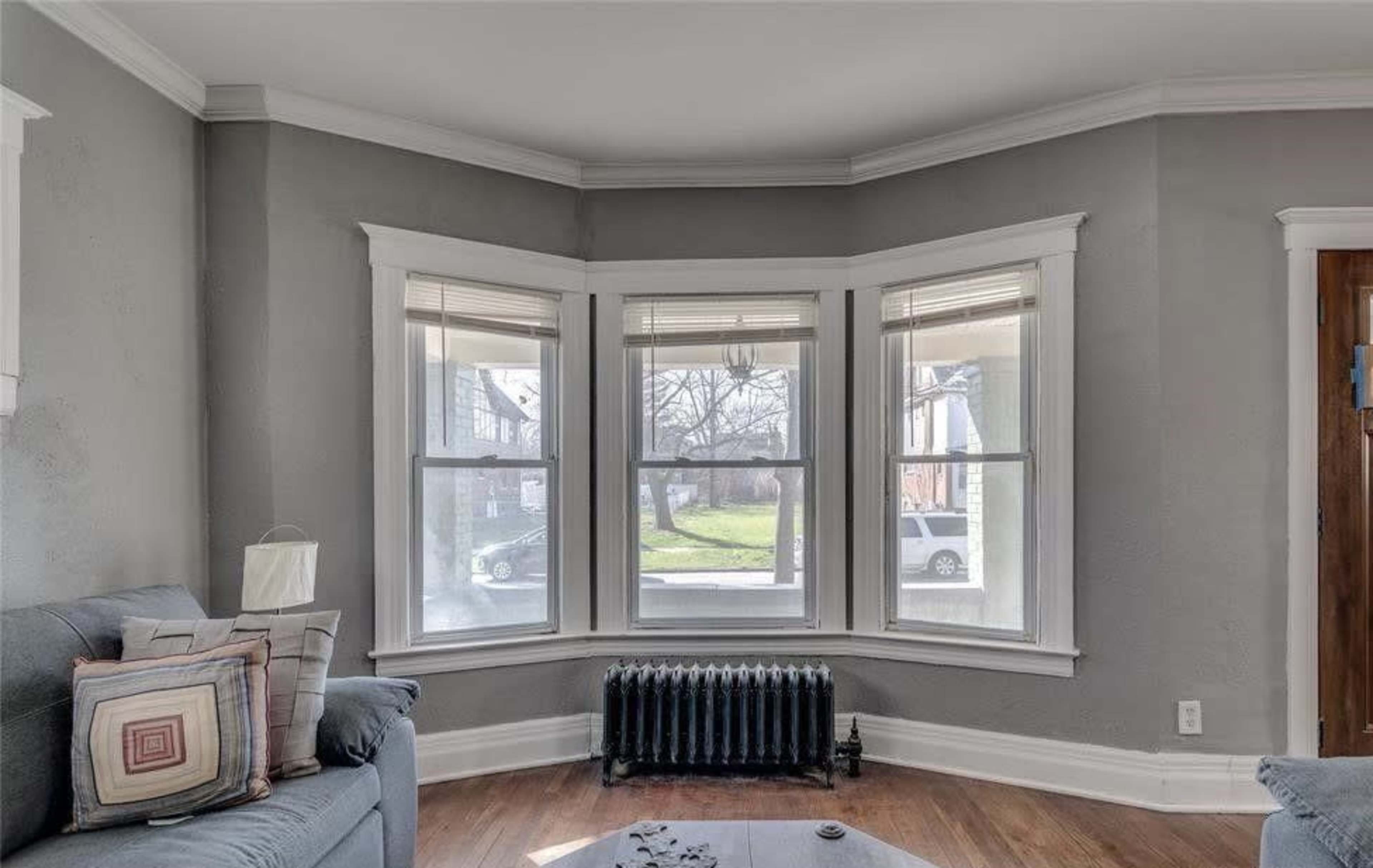The image shows a living room with a gray wall, a bay window featuring three panes, a blue sofa with decorative pillows, and a radiator beneath the window.
