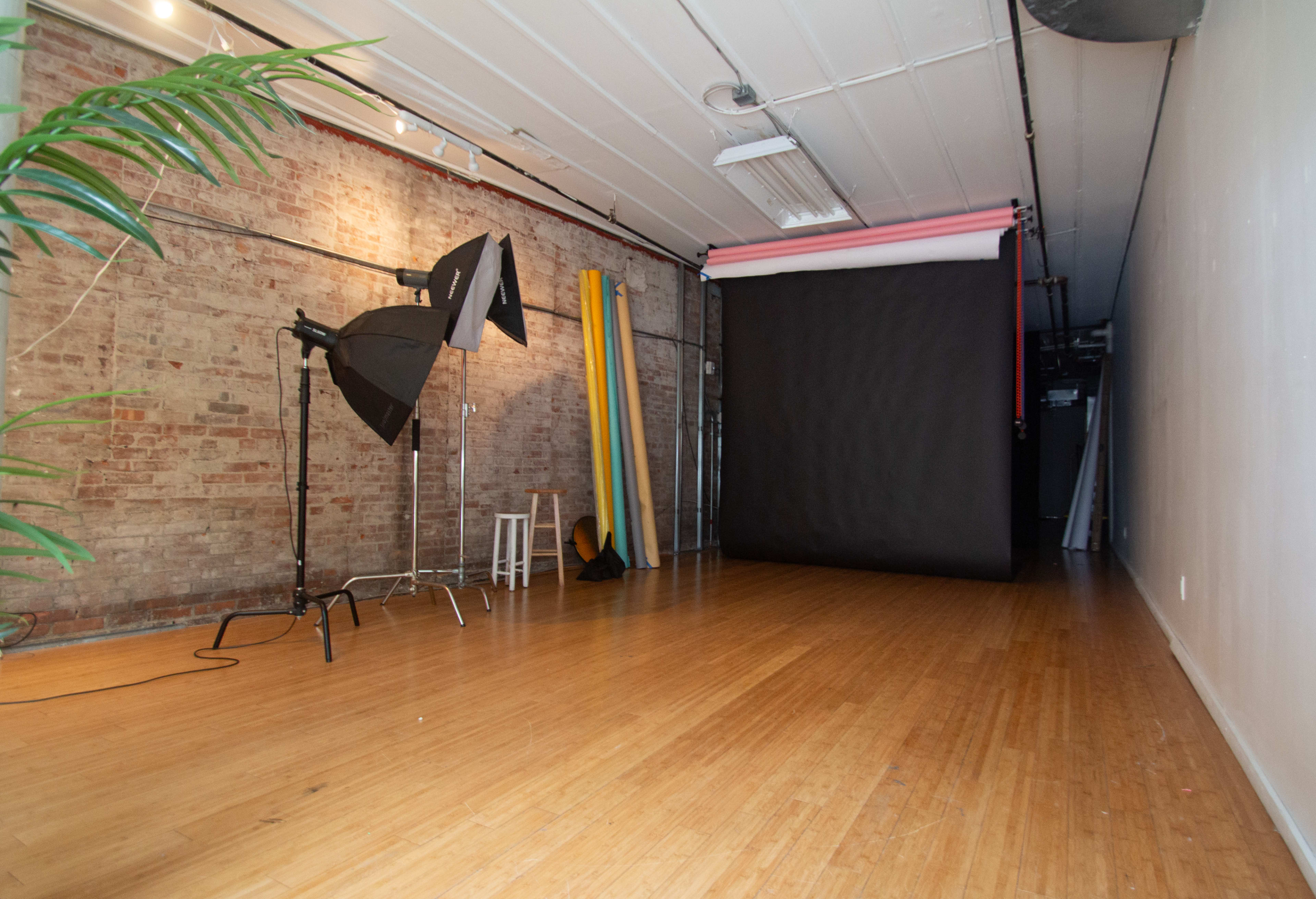 A photography studio with a wooden floor, a black backdrop, lighting equipment, and colorful rolls of paper against a brick wall.