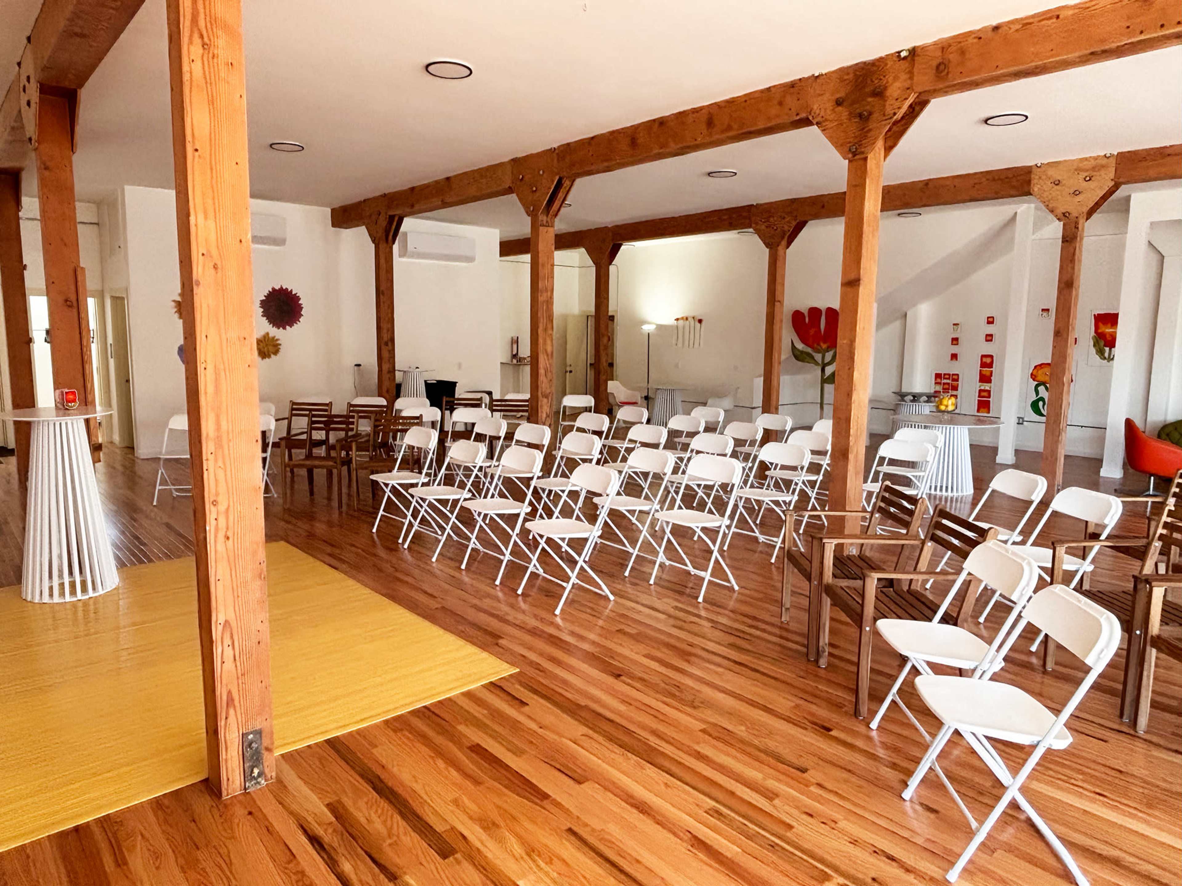 The image depicts a spacious room with wooden beams, wooden flooring, and rows of white folding chairs set up for an event.