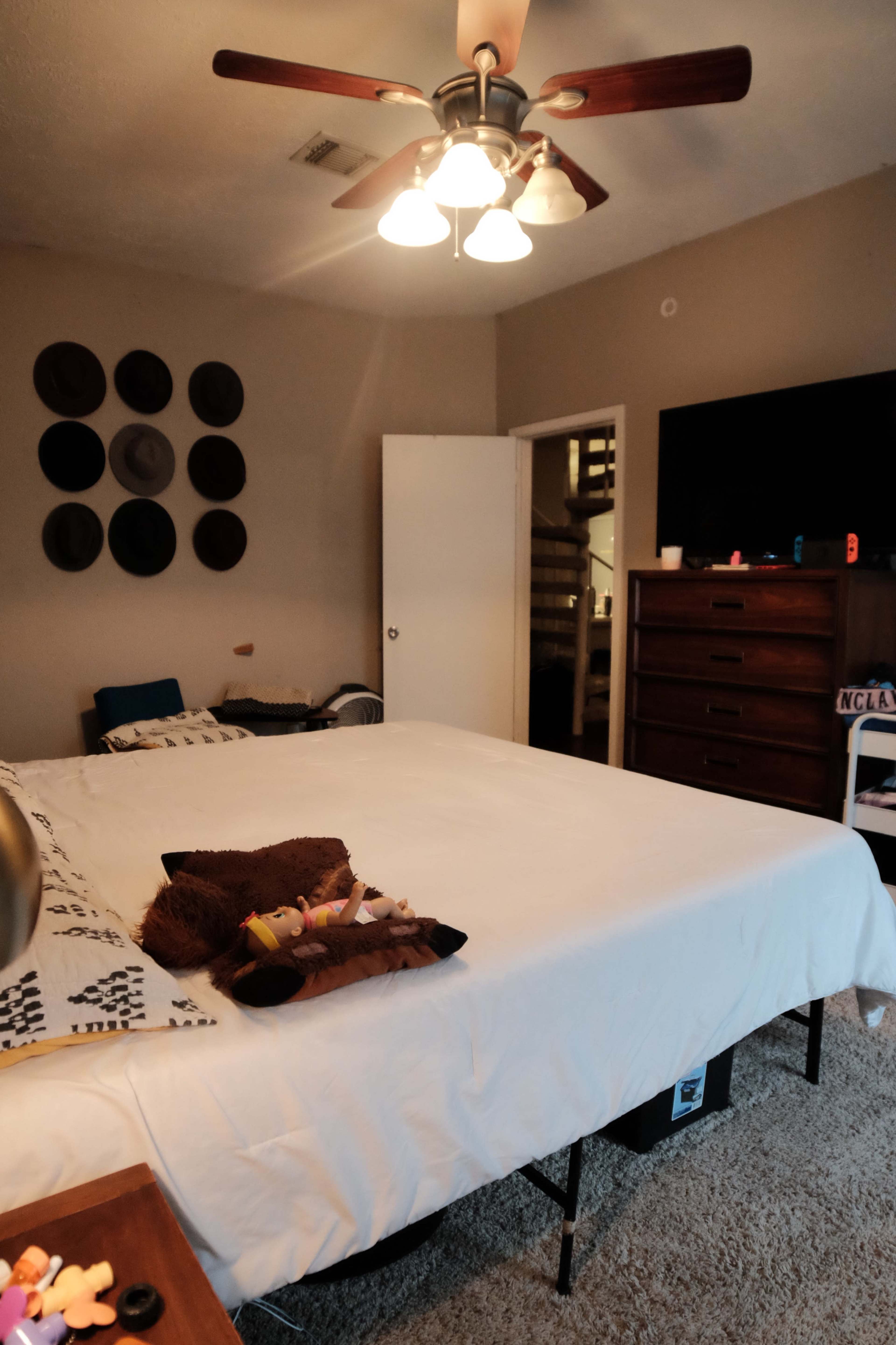 A spacious bedroom features a large bed with white bedding, a ceiling fan, and a wall adorned with various hats.