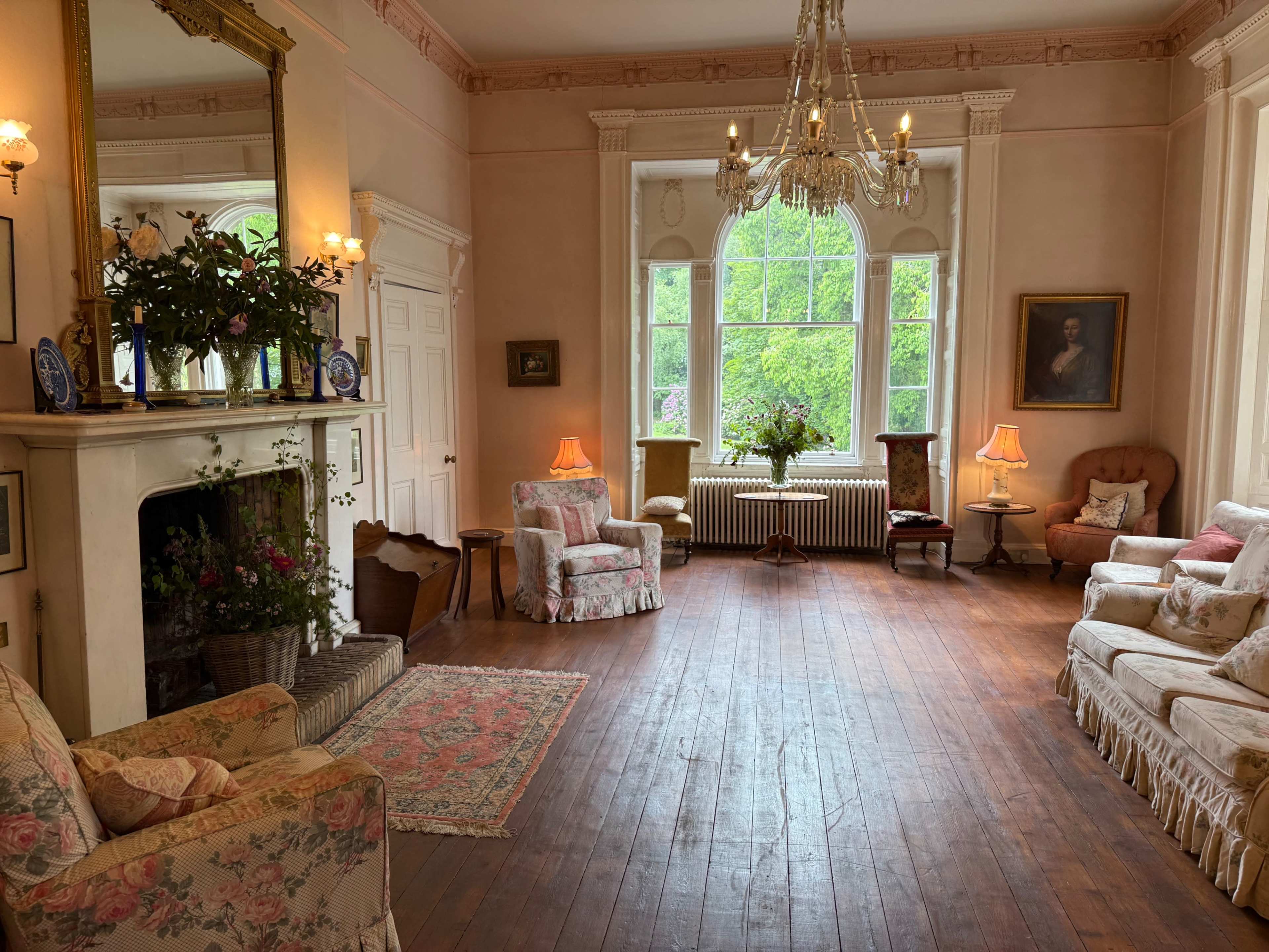 The image shows a spacious, well-lit living room with patterned armchairs, a sofa, a fireplace, and large windows overlooking greenery.
