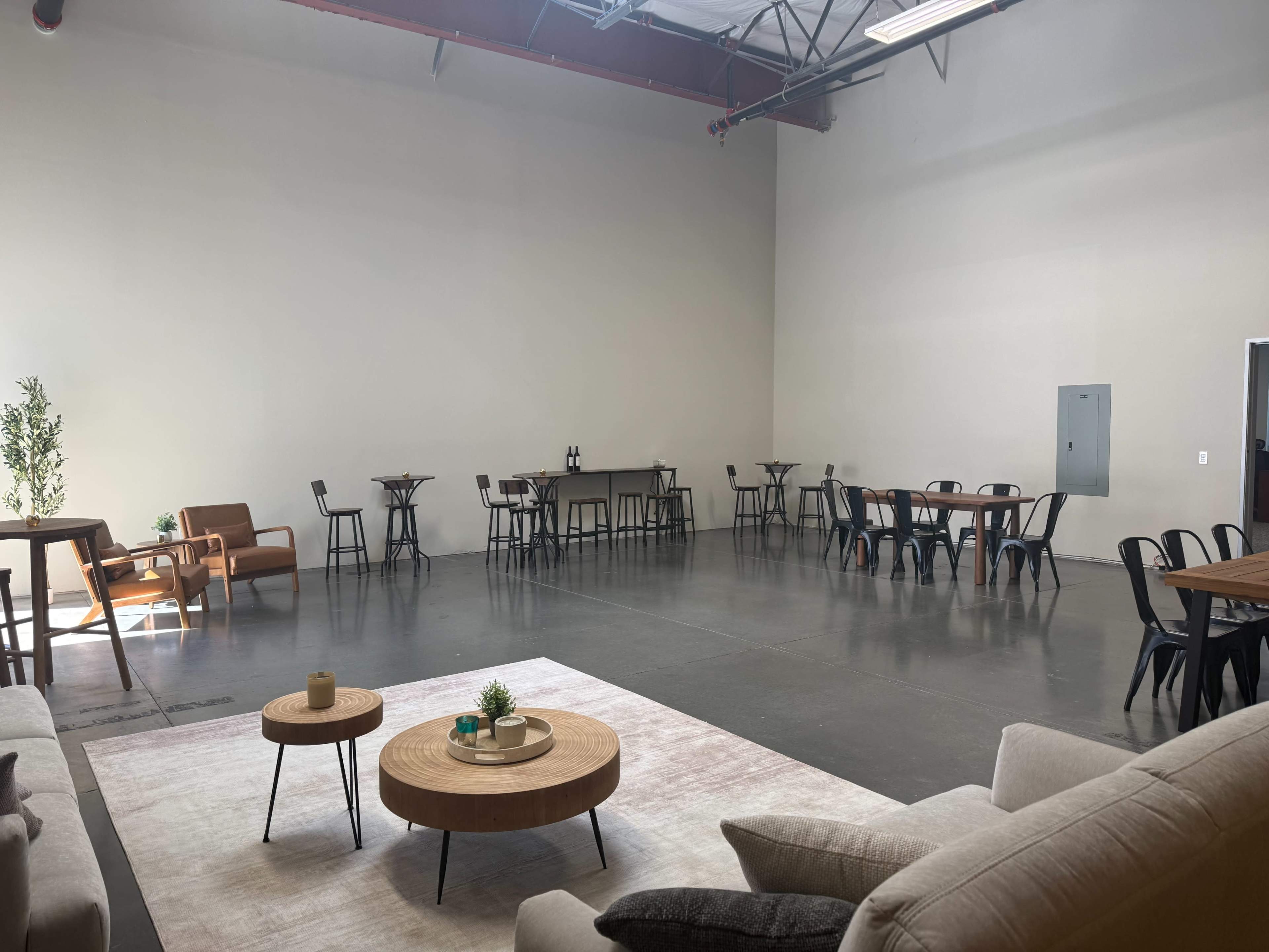 The image shows a spacious, empty room with a polished concrete floor, several tables and chairs arranged around the perimeter, and a seating area with a couch and coffee tables.