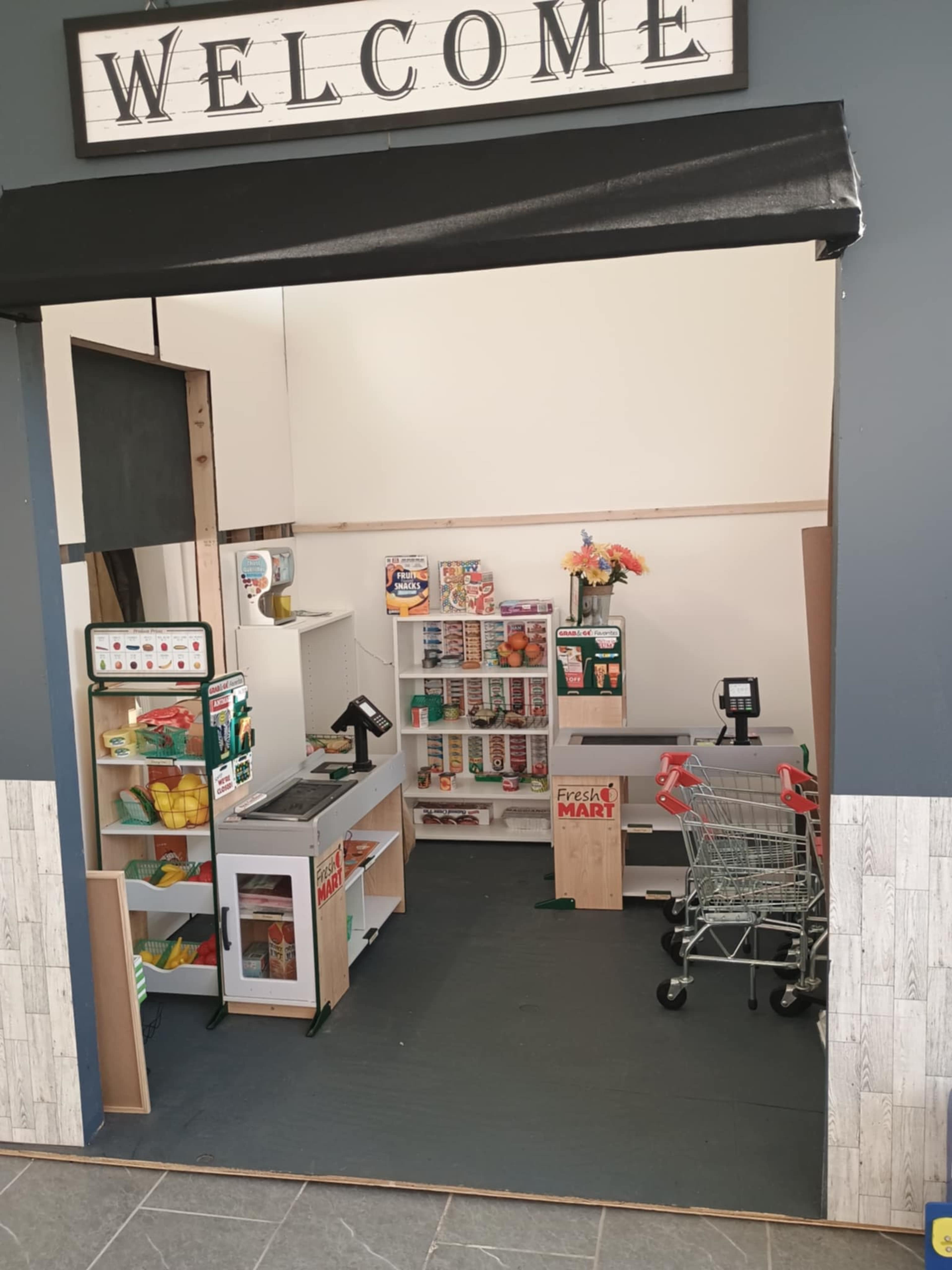 The image shows a small convenience store setup with shelves stocked with various grocery items, a checkout area, and a shopping cart.
