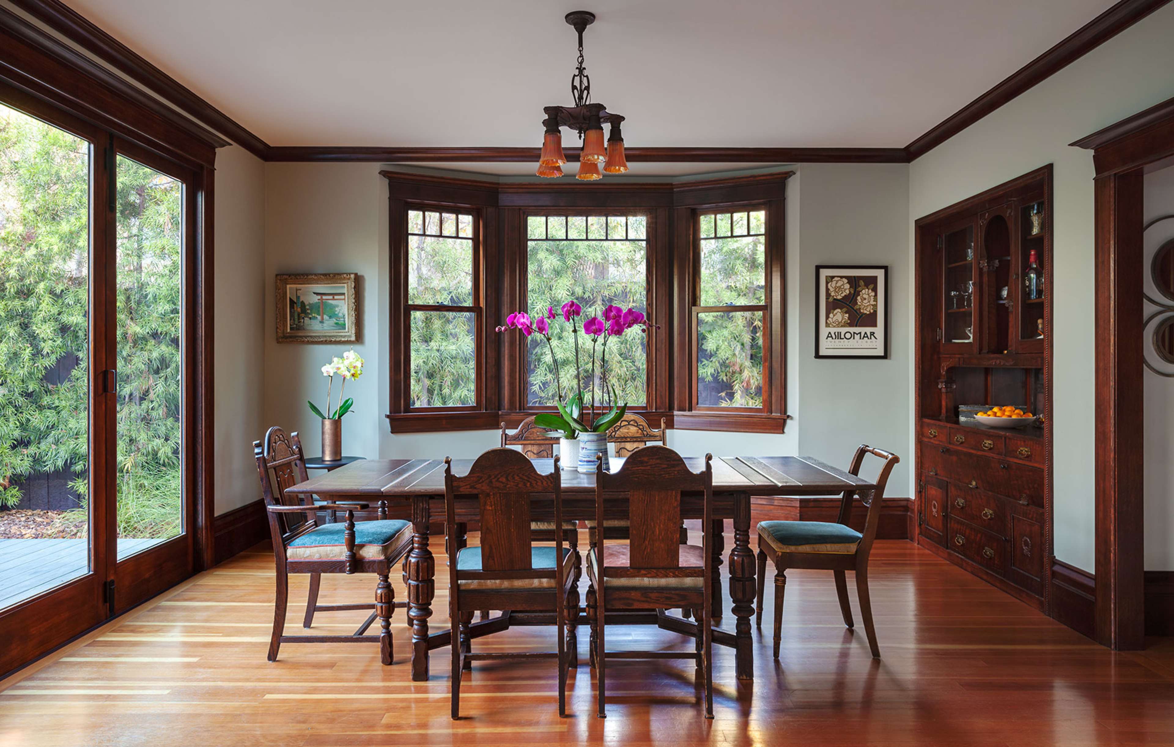 The image shows a wood-furnished dining room featuring a large table surrounded by chairs, illuminated by a pendant light and adorned with a vase of orchids.