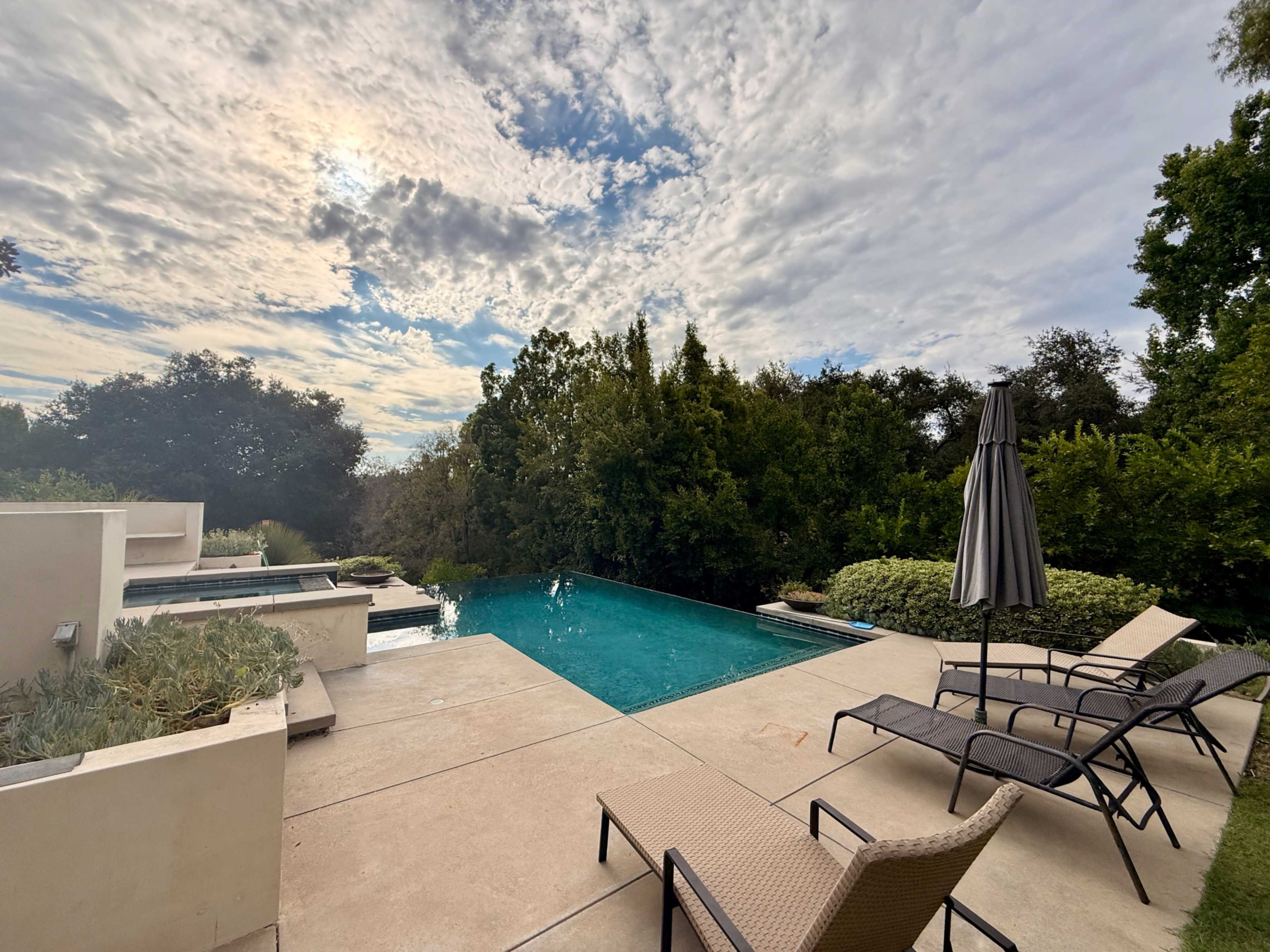 The image shows a tranquil backyard scene featuring a swimming pool, lounge chairs, and lush greenery under a cloudy sky.