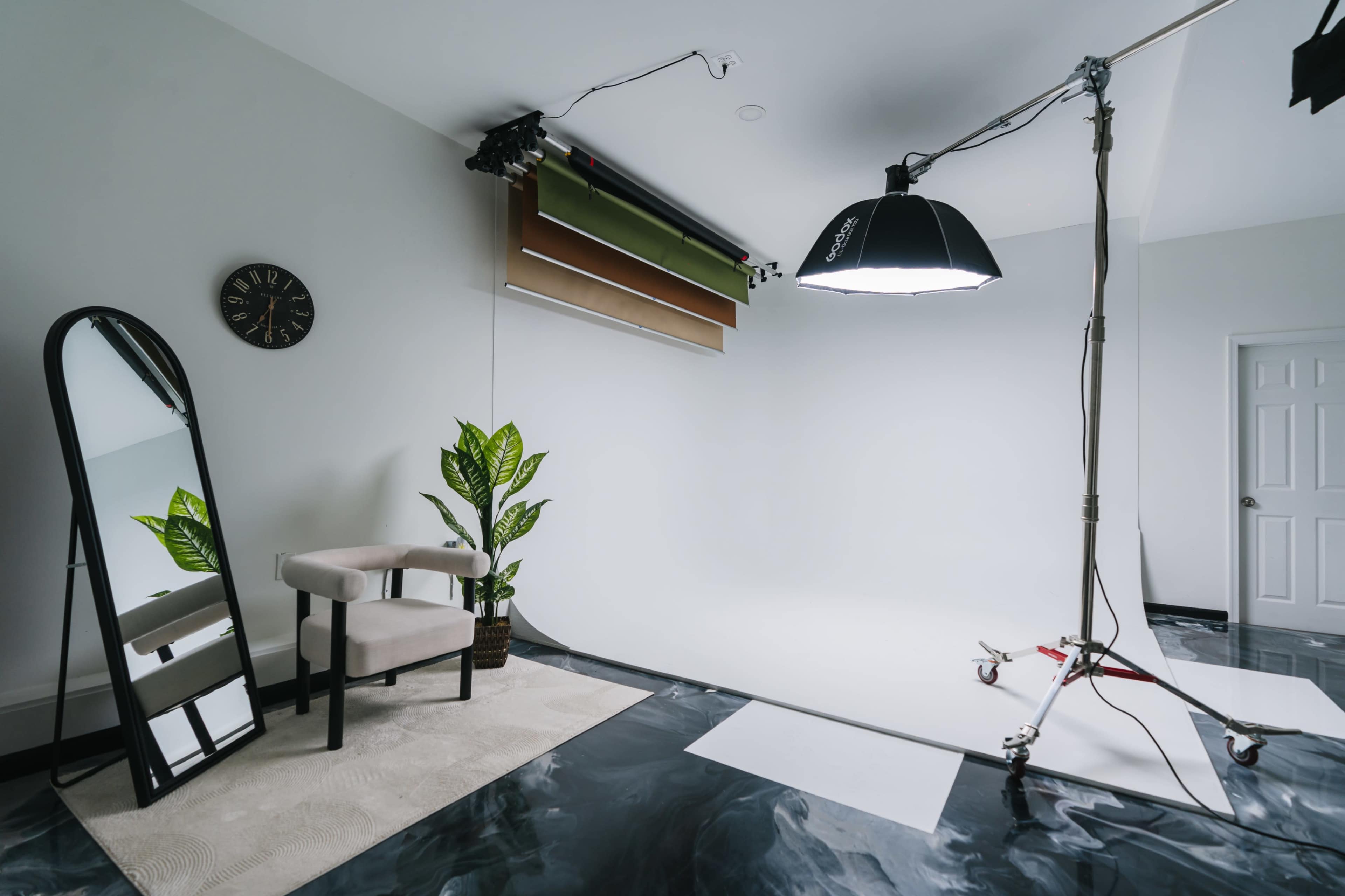 A photography studio is set up with a large light, a chair, a mirror, a potted plant, and a backdrop support system against a white wall.