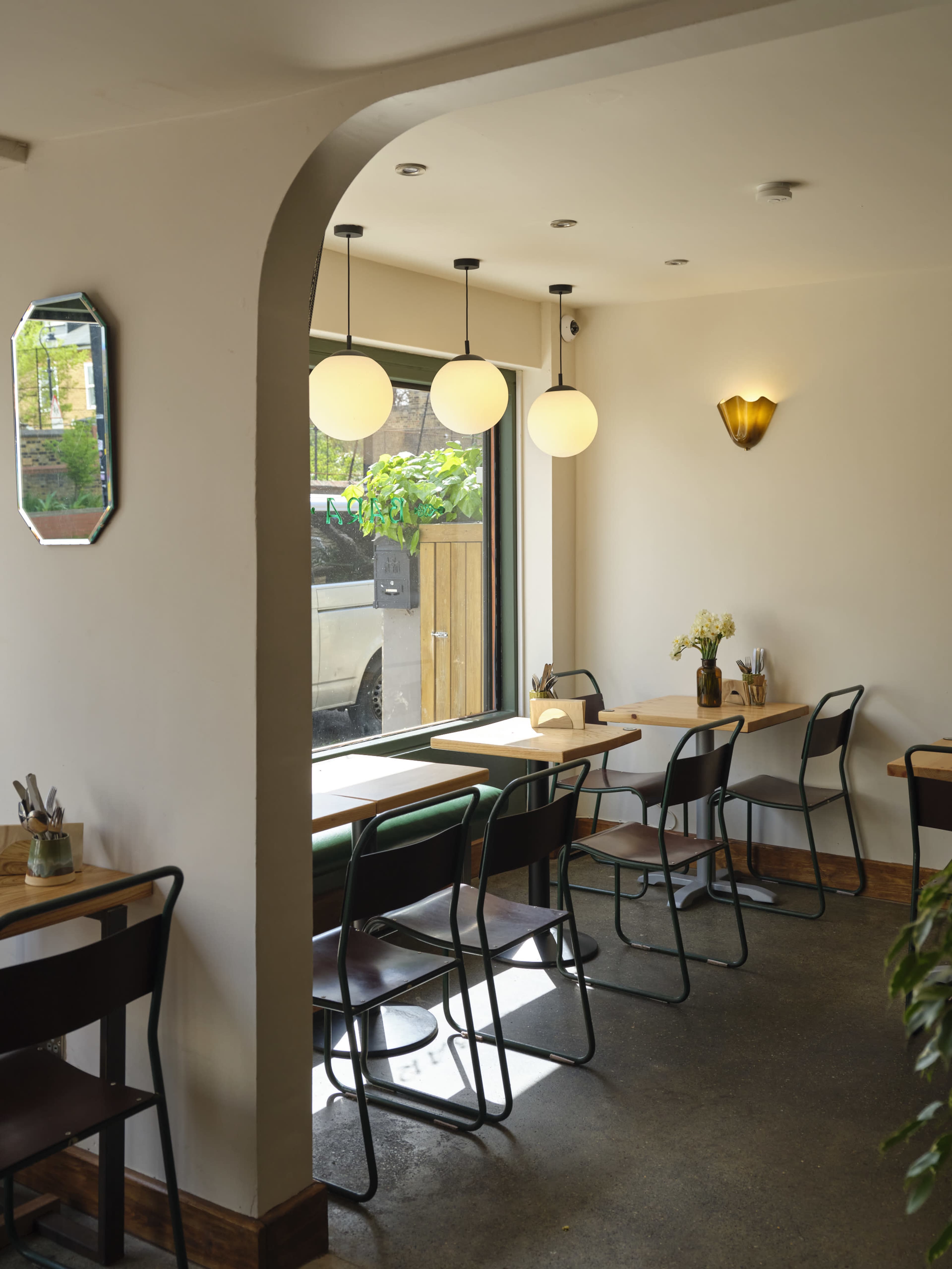 The image shows a cozy café interior with several tables and chairs arranged near large windows, illuminated by round pendant lights.