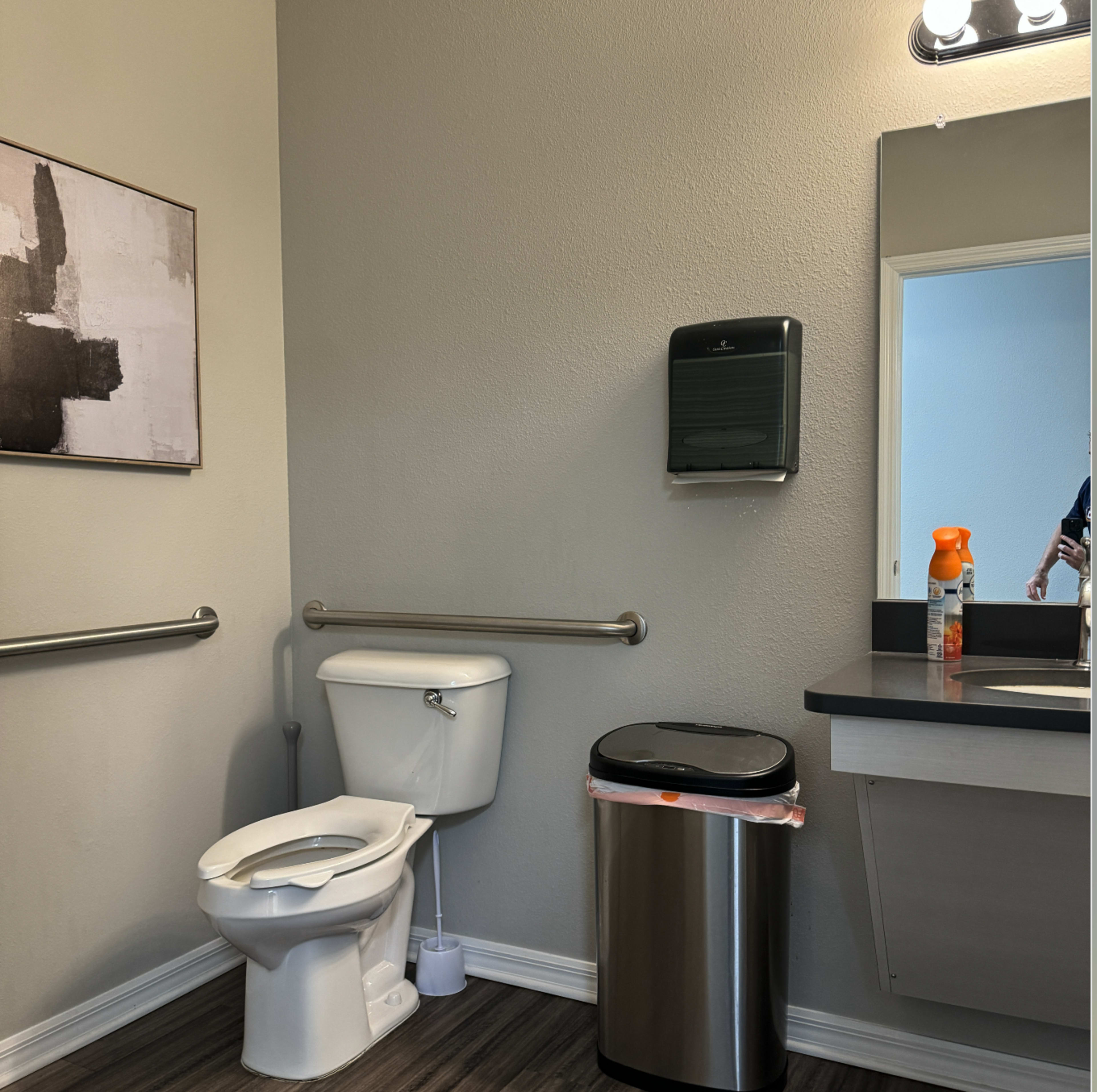 The image shows a restroom containing a toilet, a wall-mounted paper towel dispenser, a trash can, and a sink with a mirror.