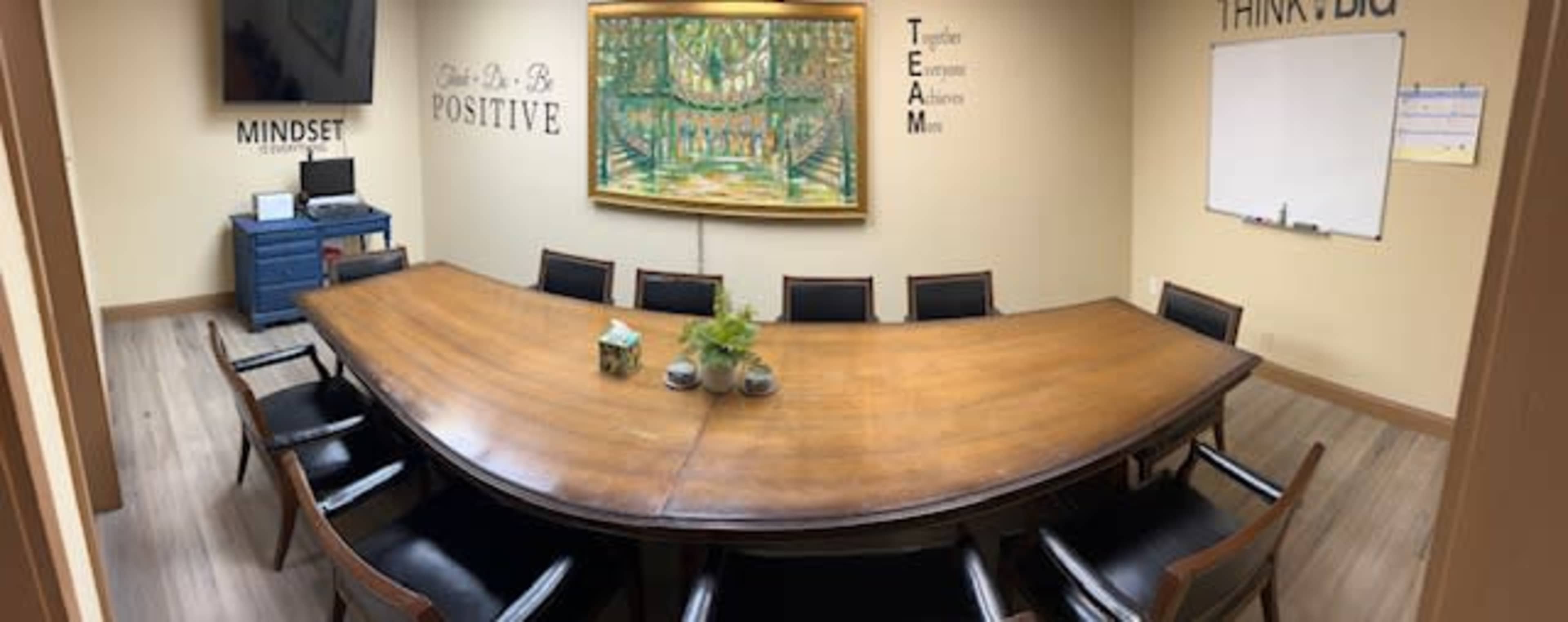 A conference room with a large wooden table surrounded by black chairs, framed by a painting and motivational words on the walls.