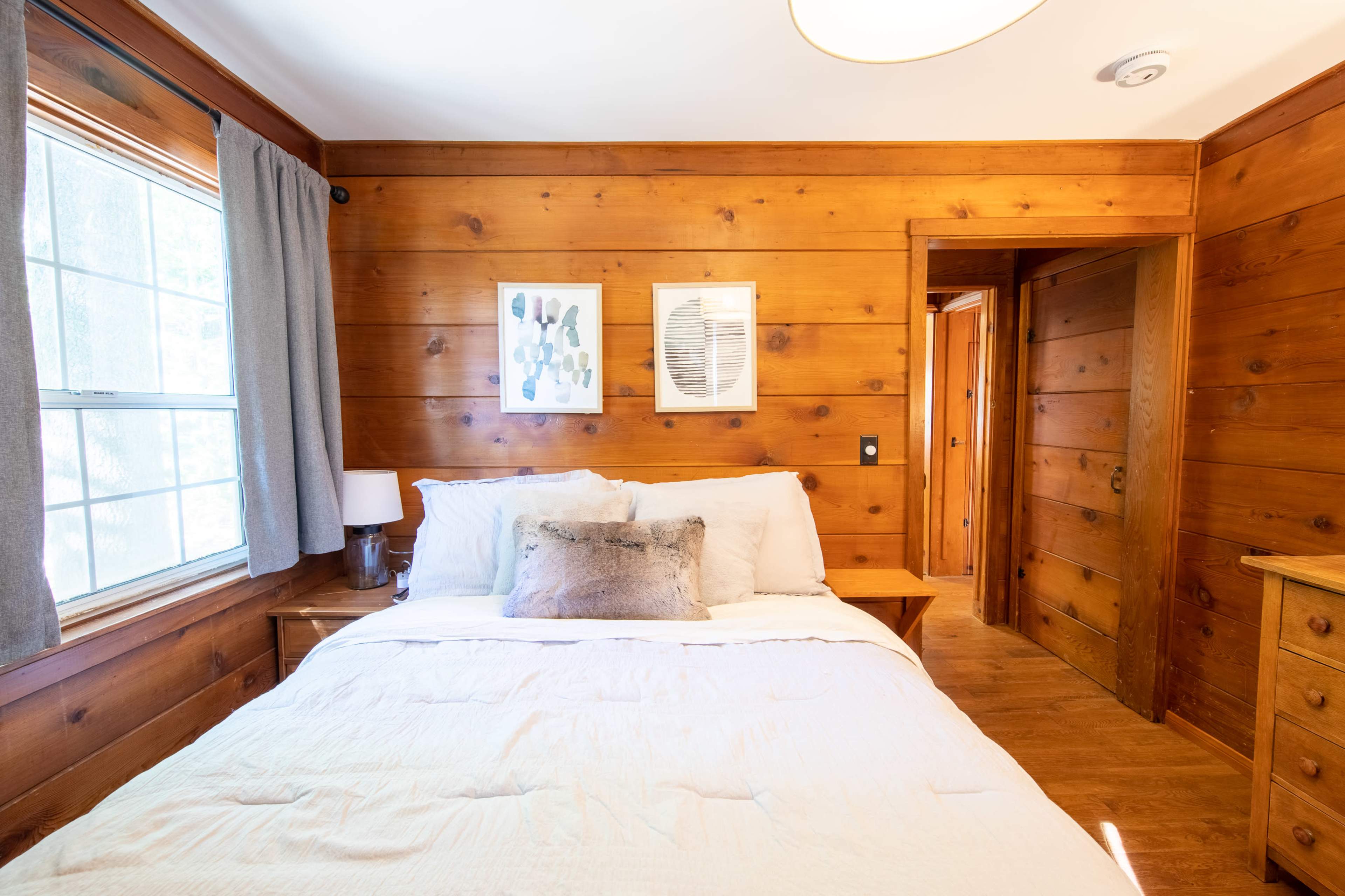 The image shows a cozy bedroom with wood-paneled walls, featuring a bed with white linens, gray curtains, and framed artwork on the walls.