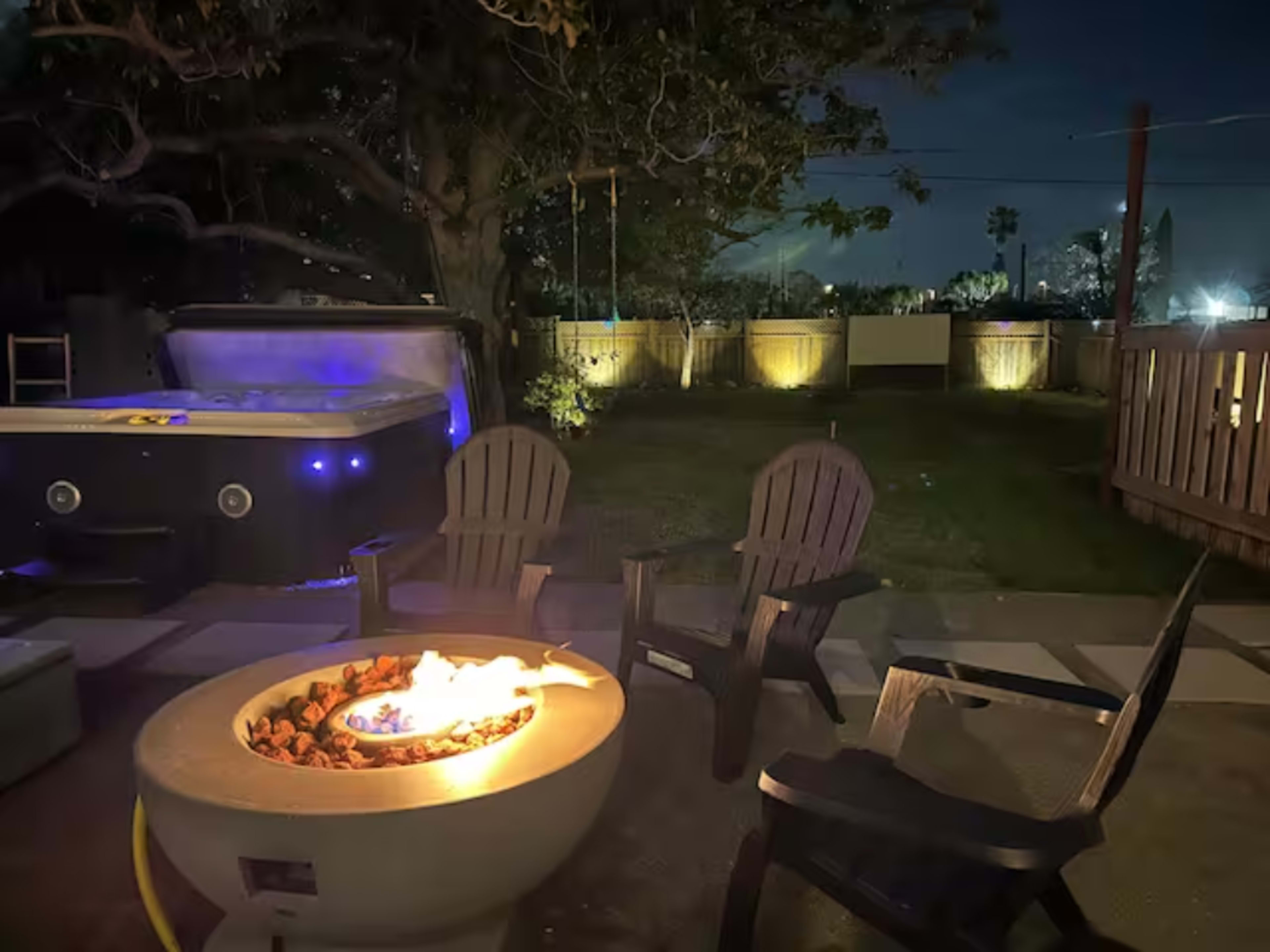 A circular fire pit surrounded by four adirondack chairs is lit in a backyard at night, with a hot tub and trees in the background.