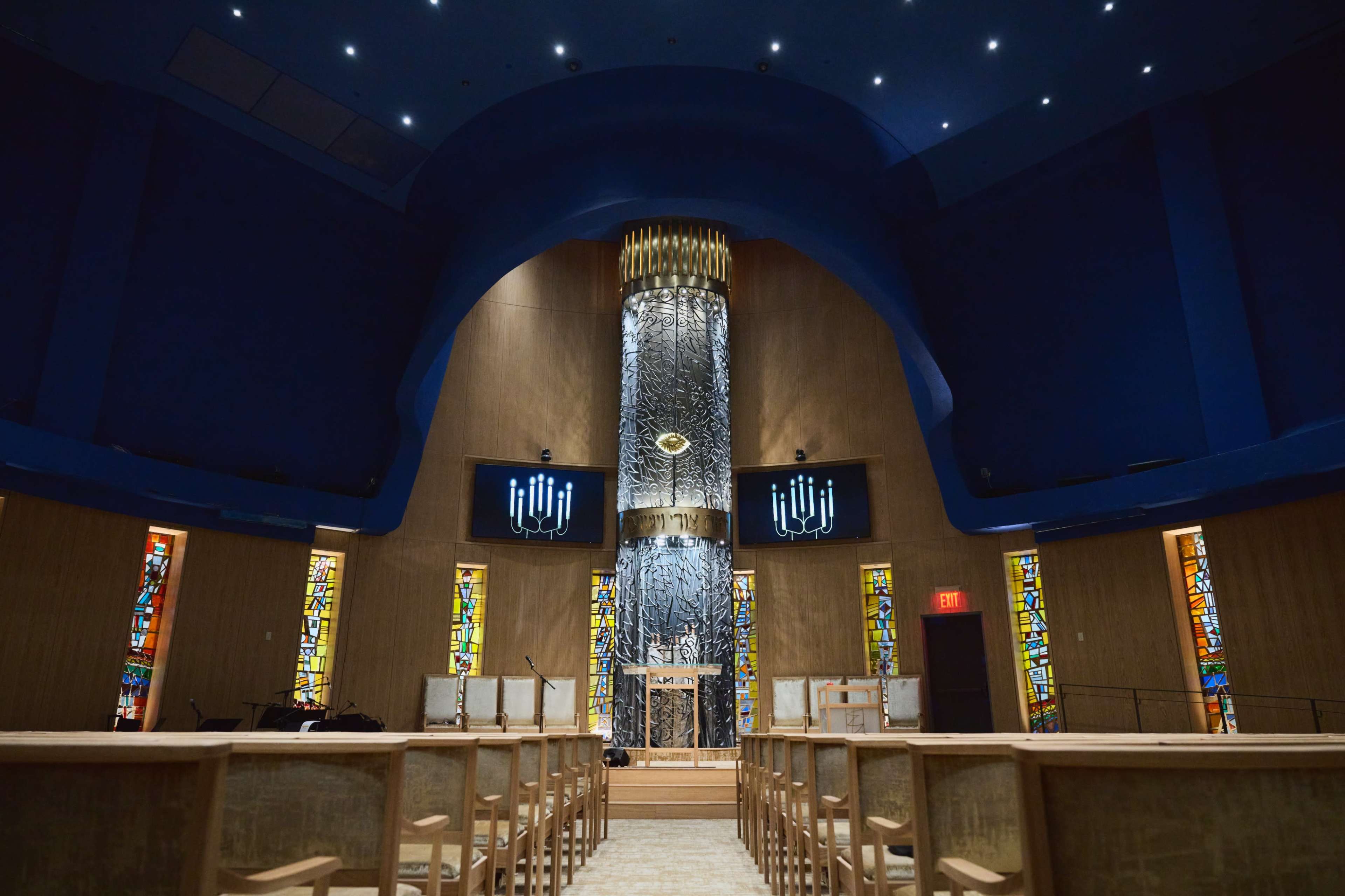 The interior of a synagogue features a high, arched ceiling, a central bema with a decorative backdrop, and stained glass windows on either side.