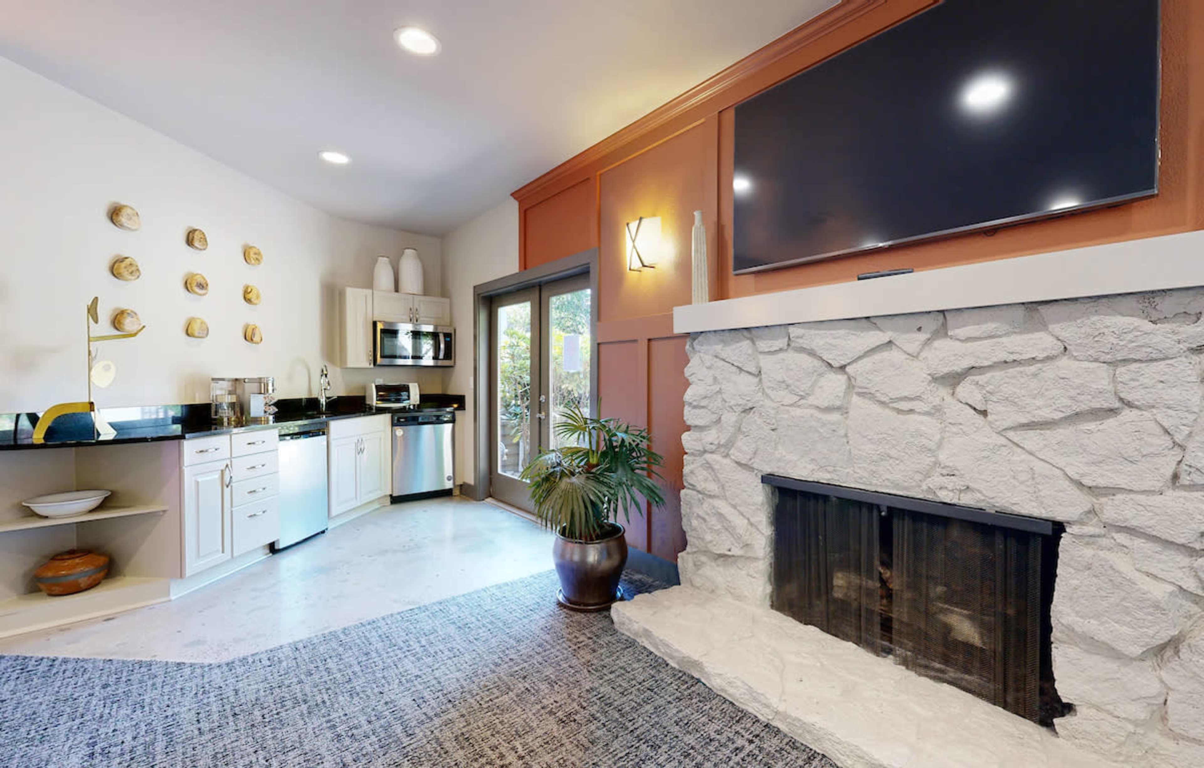 A modern living area with a stone fireplace, a kitchen area, and large windows leading to an outdoor space.