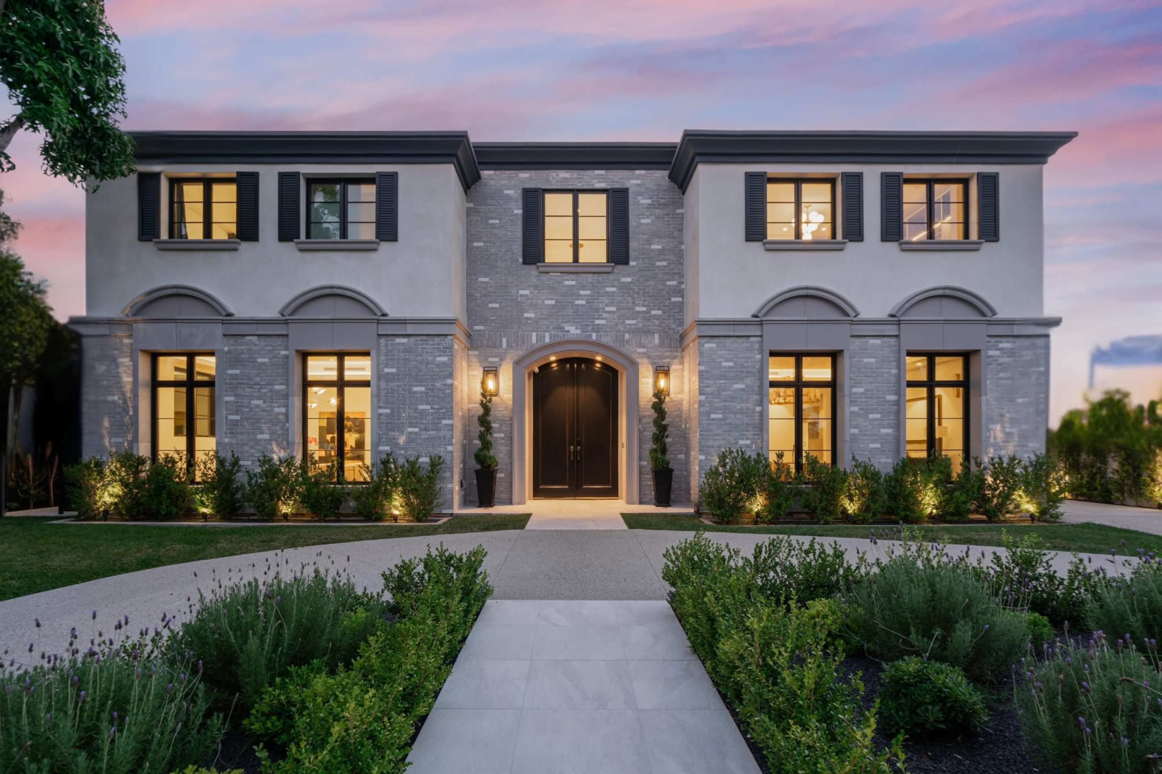The image depicts a modern two-story home with a brick facade, large windows, and symmetrical design, surrounded by landscaped greenery.
