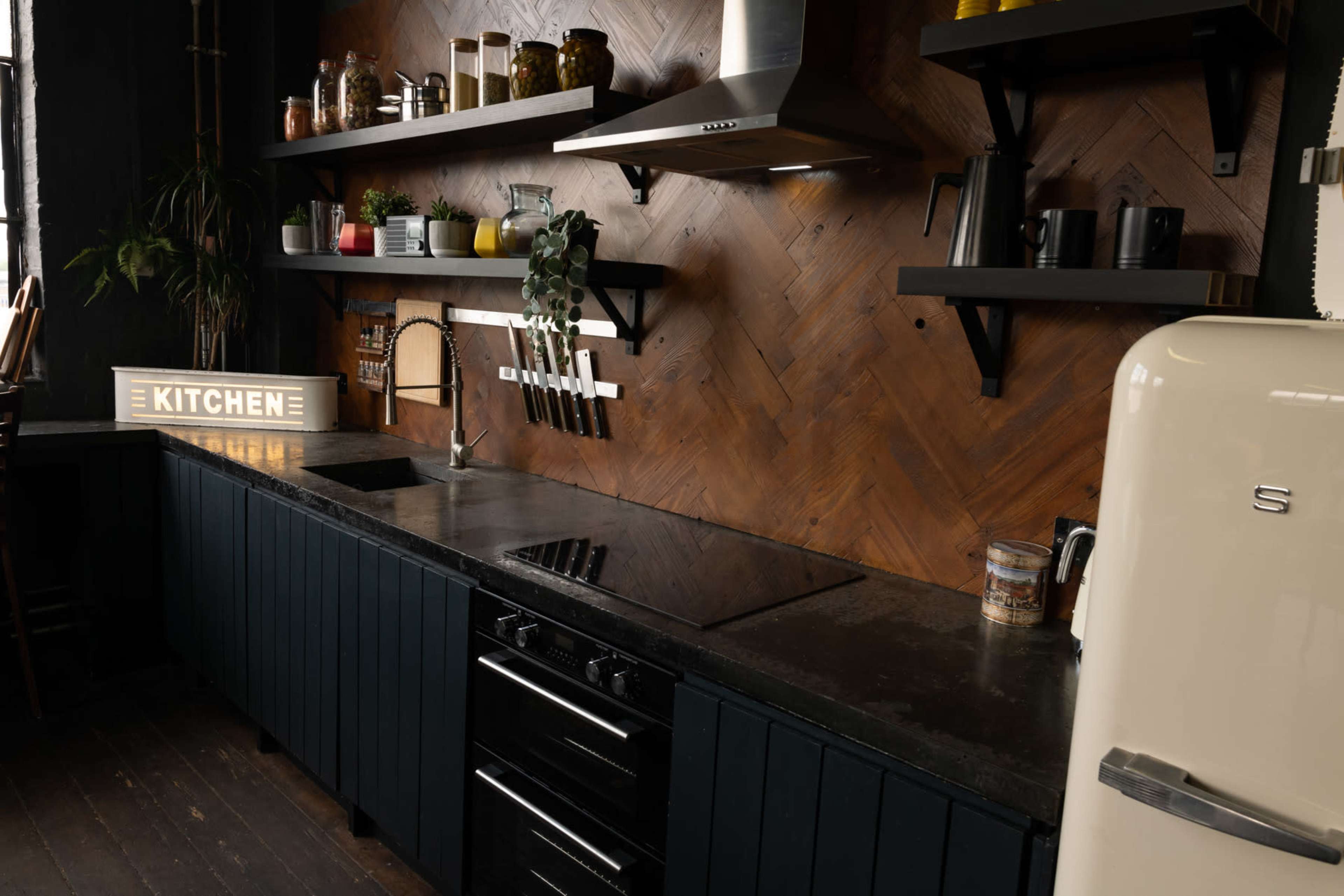 A modern kitchen with a wooden herringbone wall, black countertops, and stainless steel appliances, including a stovetop and a retro-style refrigerator.