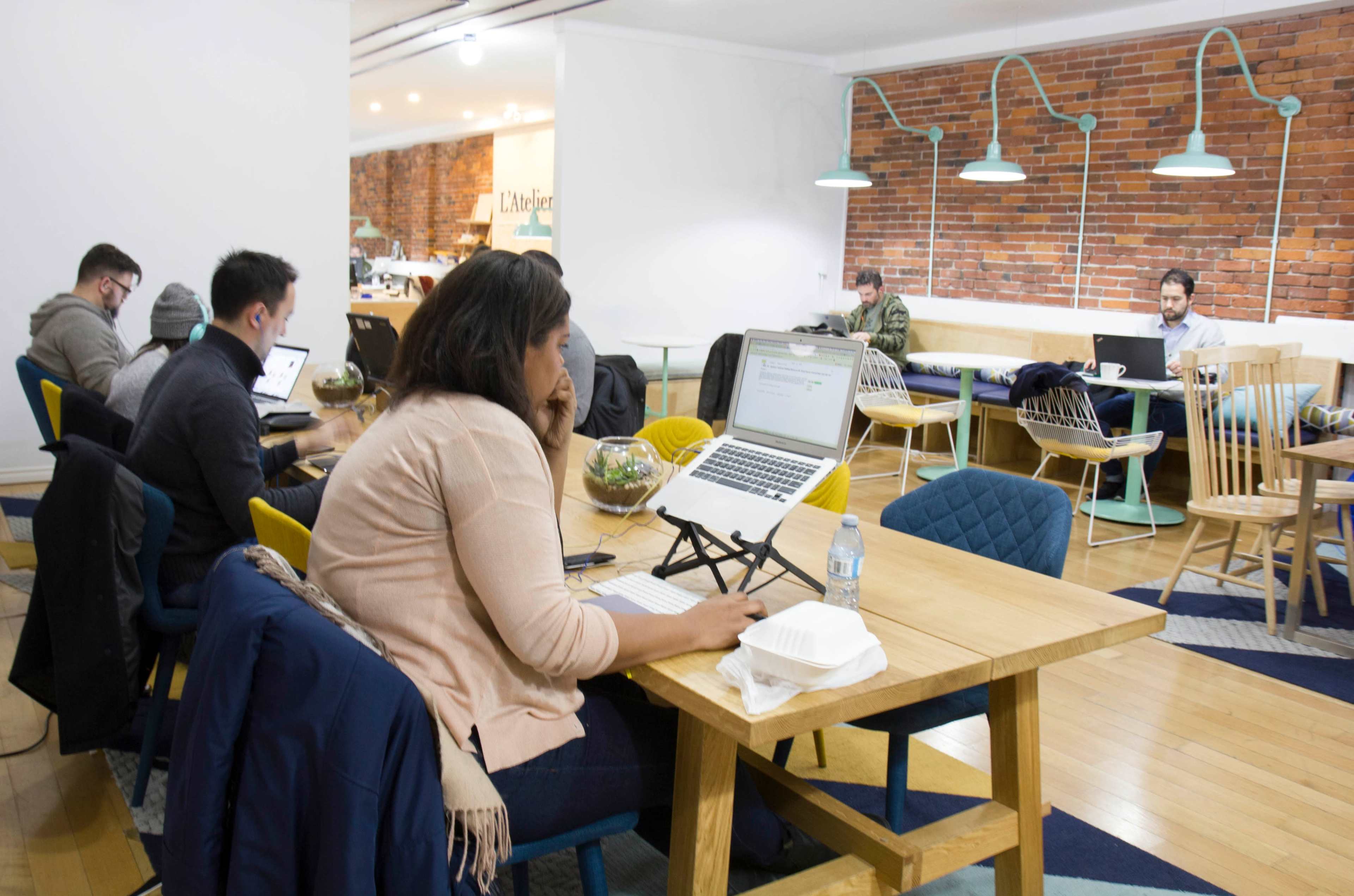 A group of people is working on laptops in a spacious, modern co-working space with wooden tables and brick walls.