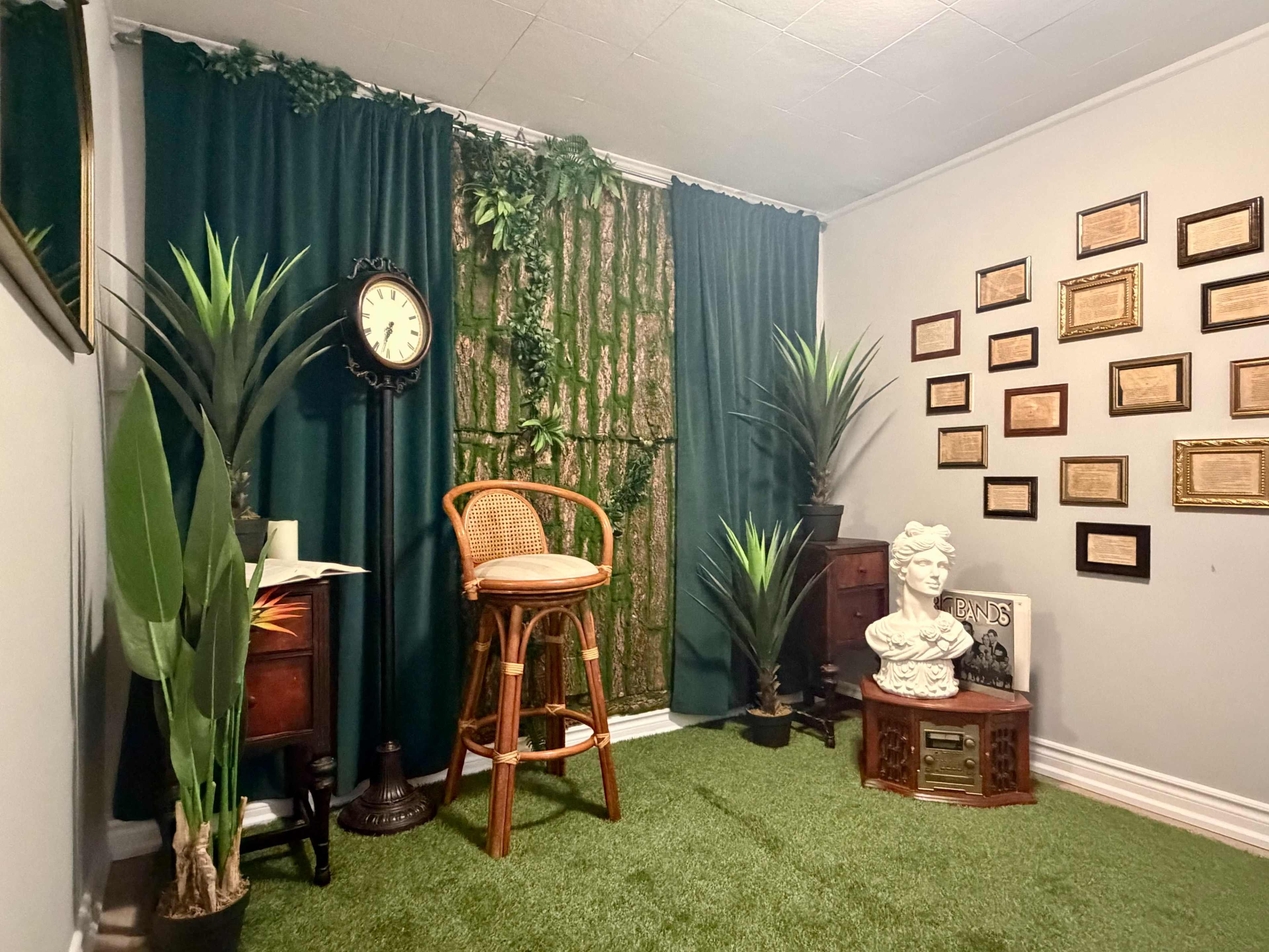 The image shows a room with green curtains and artificial grass flooring, featuring a clock on a pedestal, a bar stool, plants, and framed plaques on the walls.