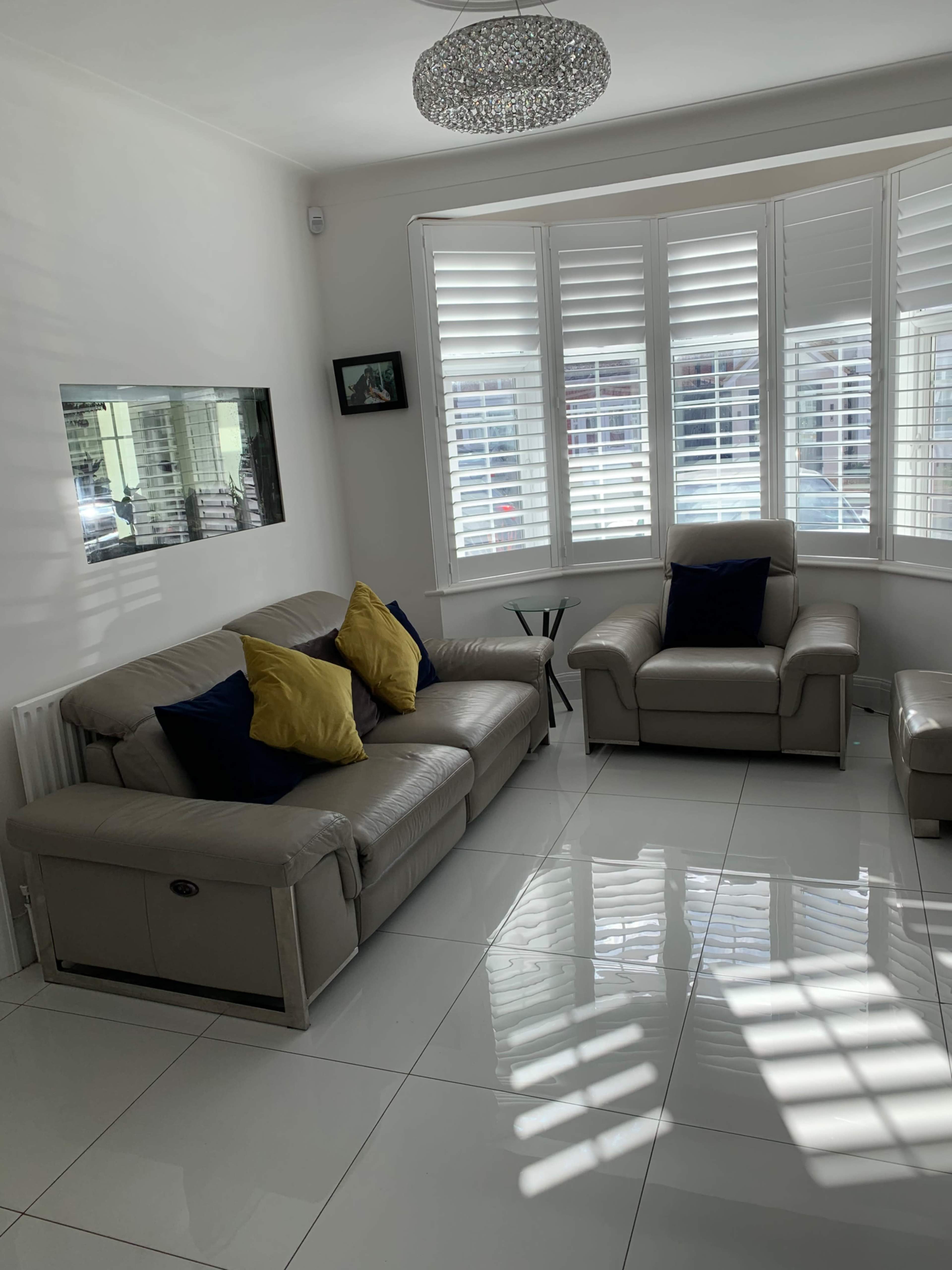 The living room features a light-colored leather sofa and armchair, a glass side table, and plantation shutters allowing natural light to create patterns on the tiled floor.