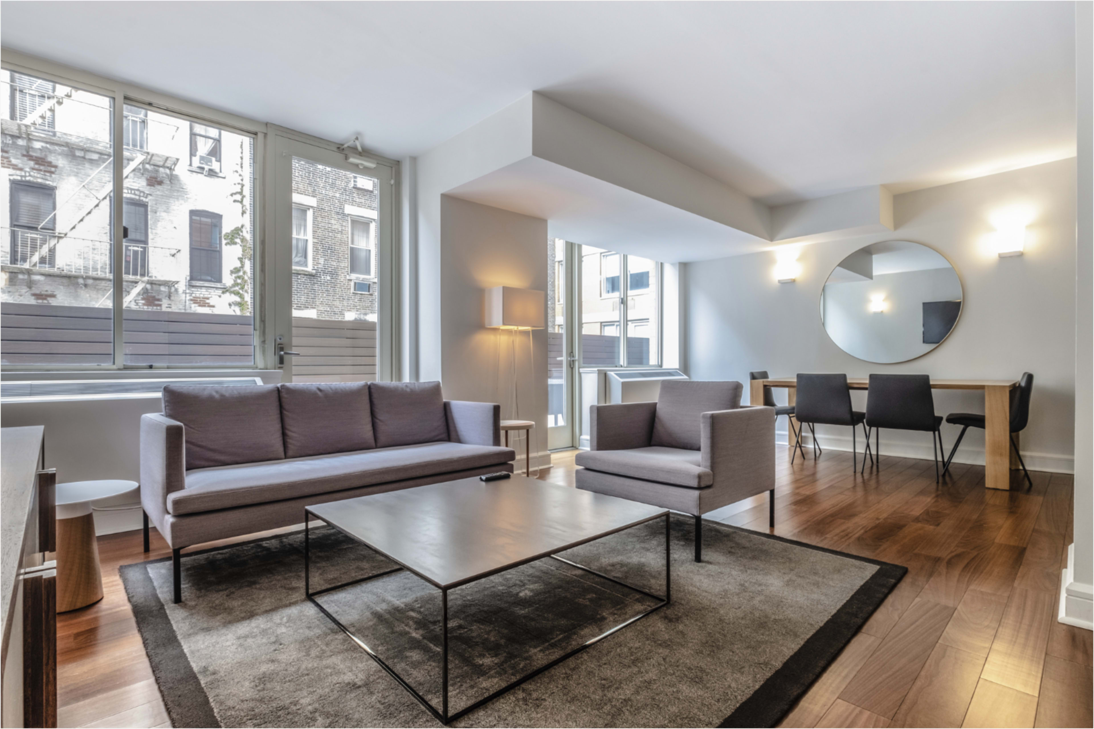 The image shows a modern living room featuring two gray sofas, a glass coffee table, and a dining area with a wooden table and black chairs, all in a bright space with large windows.