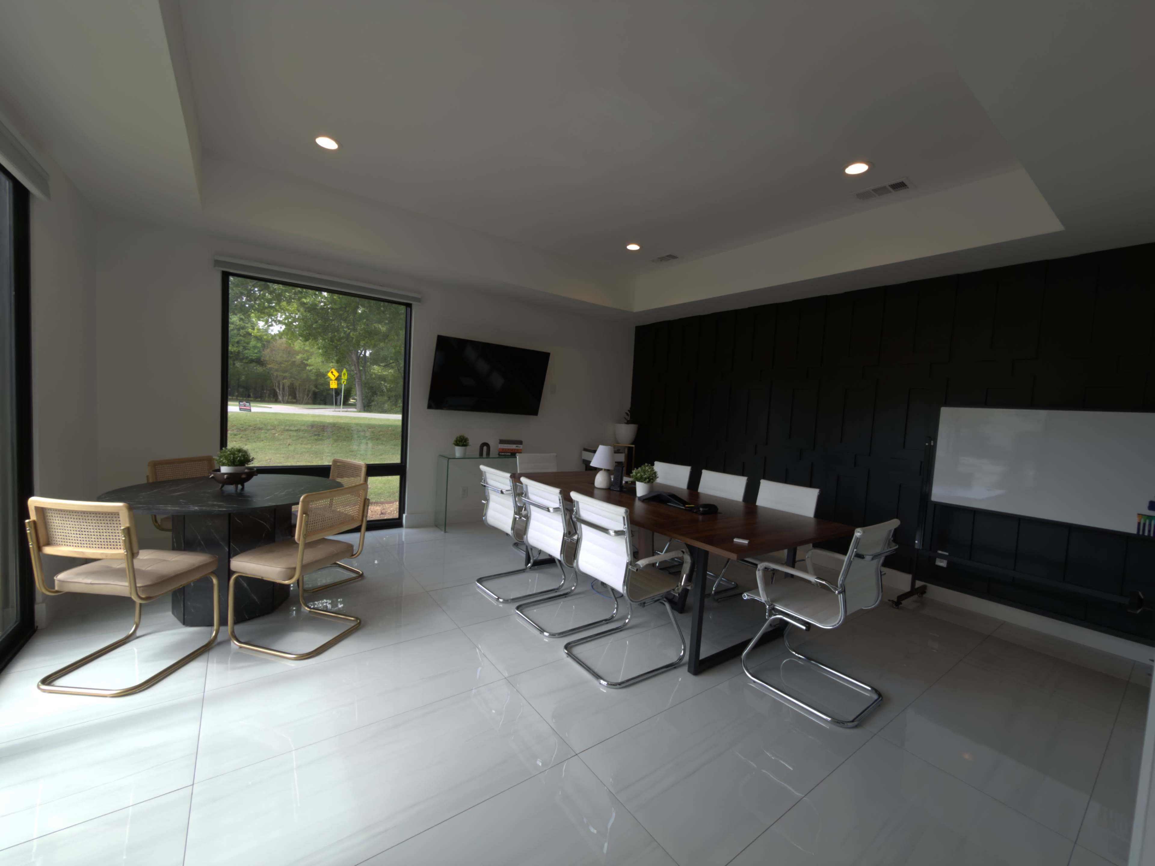 The image shows a modern conference room with a large table surrounded by white chairs, a flat-screen TV on the wall, and large windows overlooking a green area.