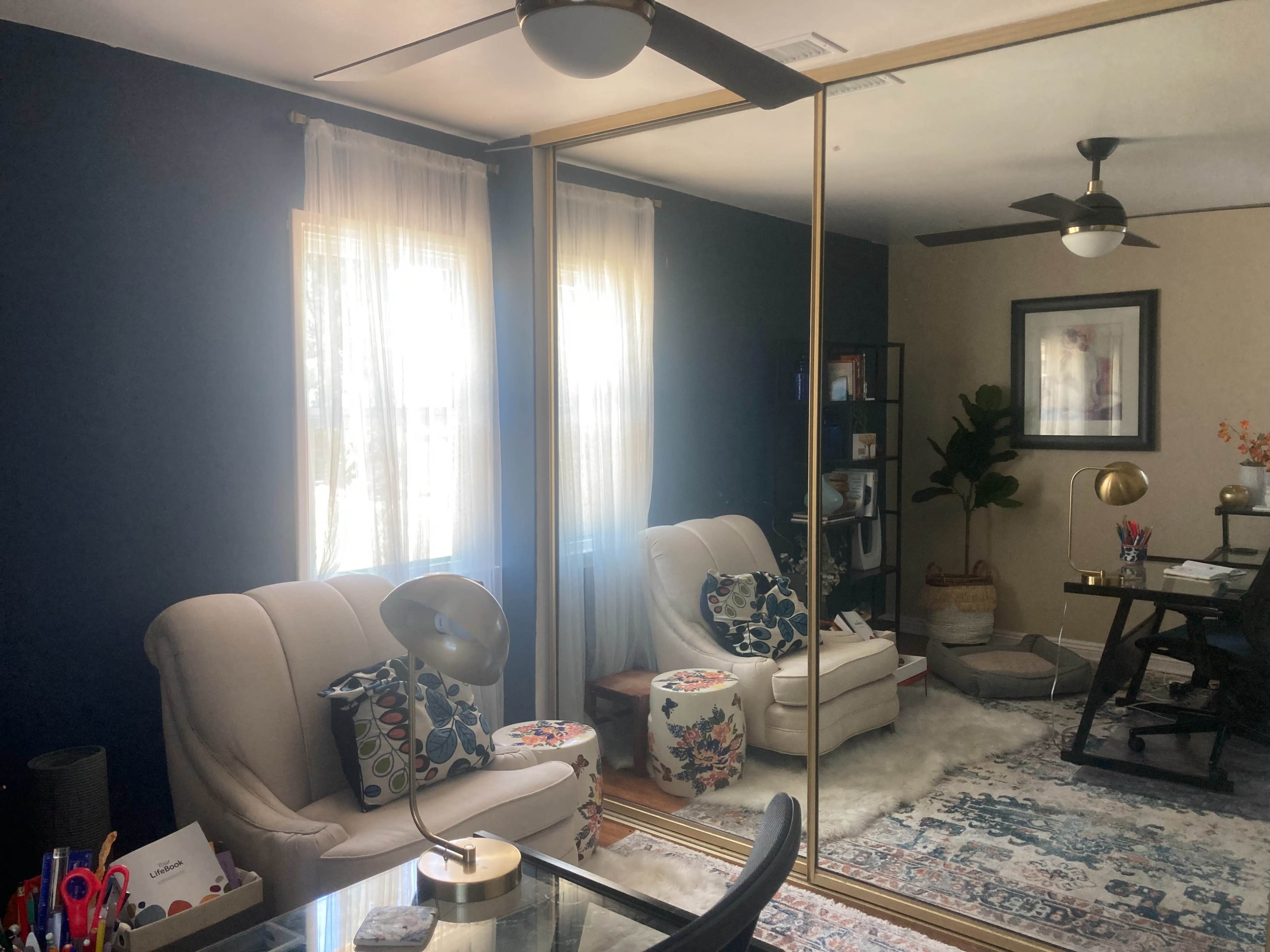 The room features a seating area with a pair of white chairs, a desk, and floor-to-ceiling mirrored closets reflecting natural light from the windows.