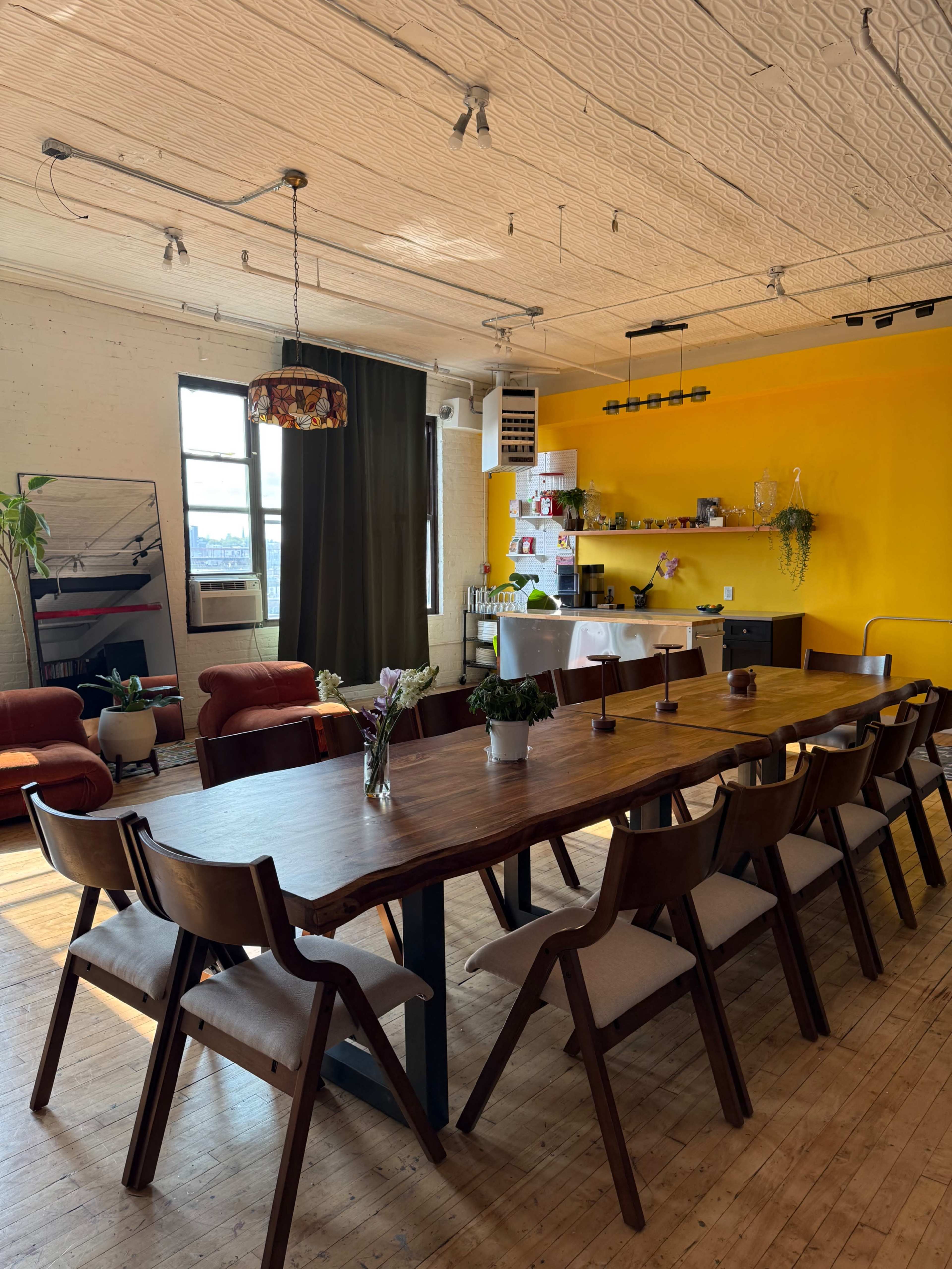 A spacious dining area features a long wooden table surrounded by chairs, with a yellow accent wall and a kitchen area in the background.