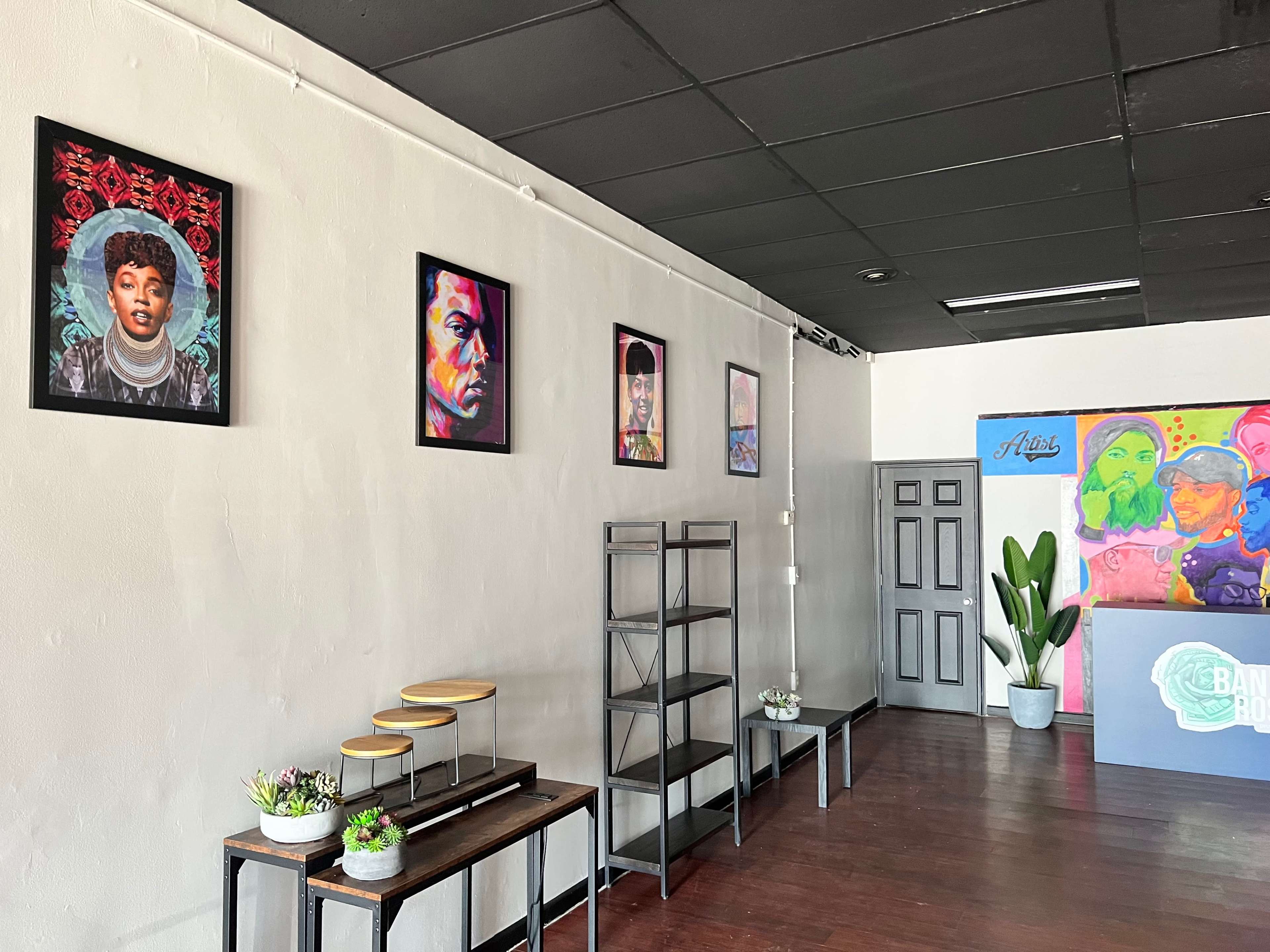A modern interior space featuring framed portraits on a gray wall, a set of wooden shelves, and a reception area with a colorful mural.