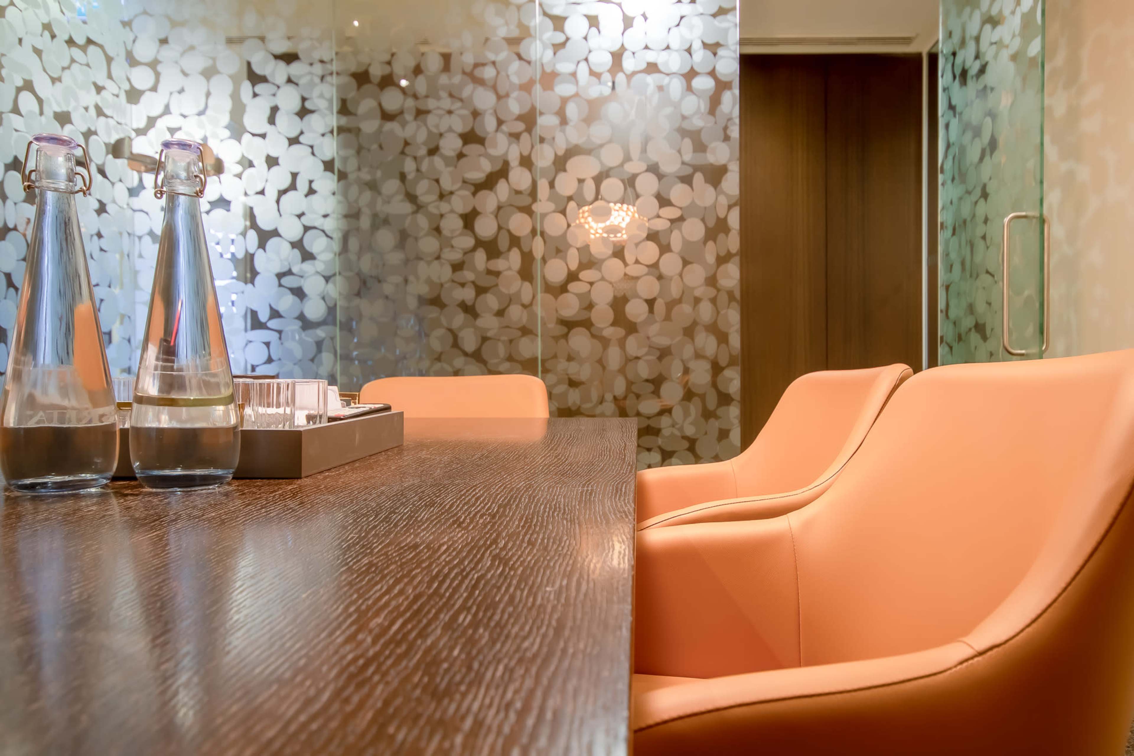 A conference room features a wooden table with two glass water bottles and modern orange chairs, set against a decorative frosted glass wall.