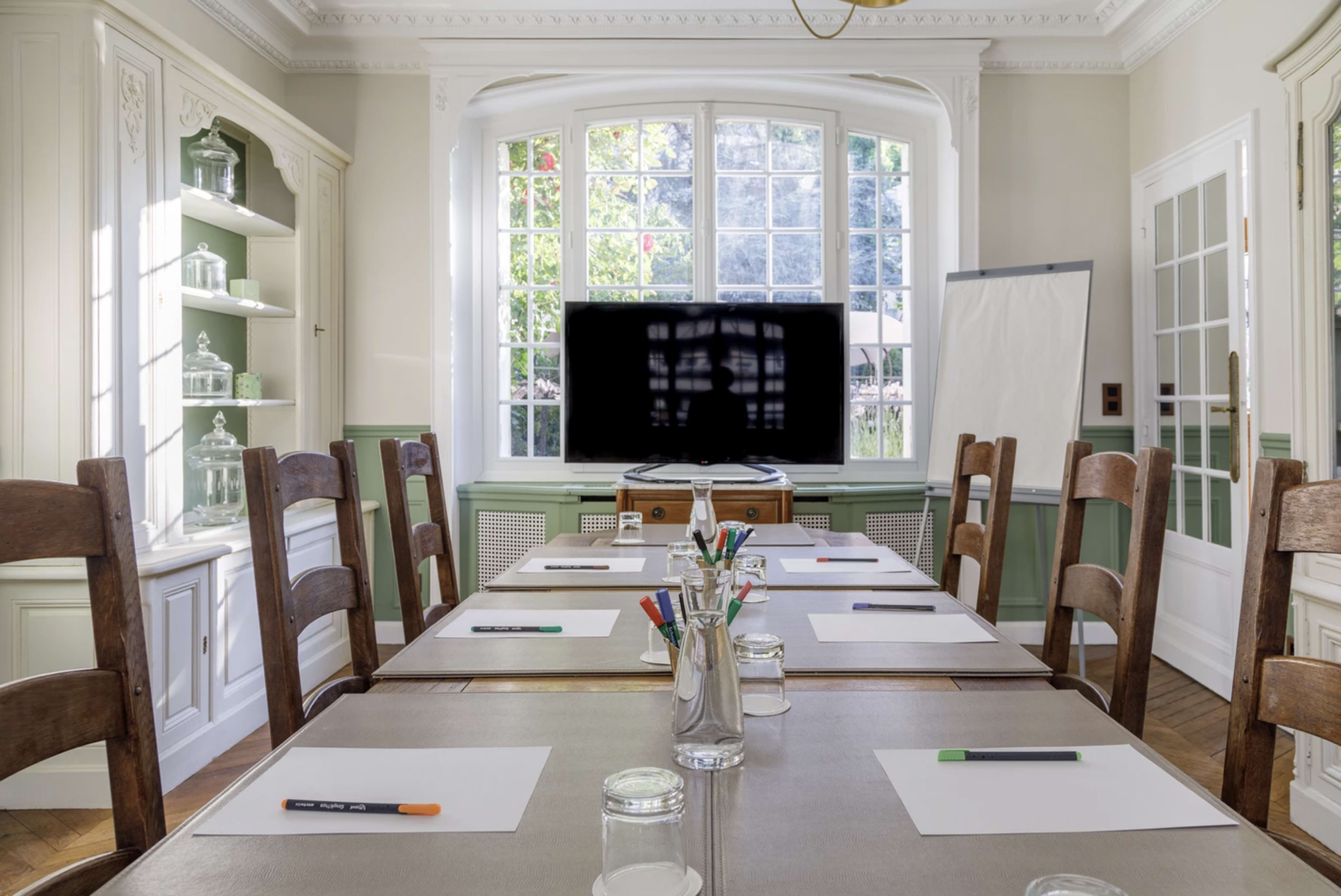 A conference room is set up with a large table, surrounded by wooden chairs, and features a window allowing natural light, a television, and a whiteboard.