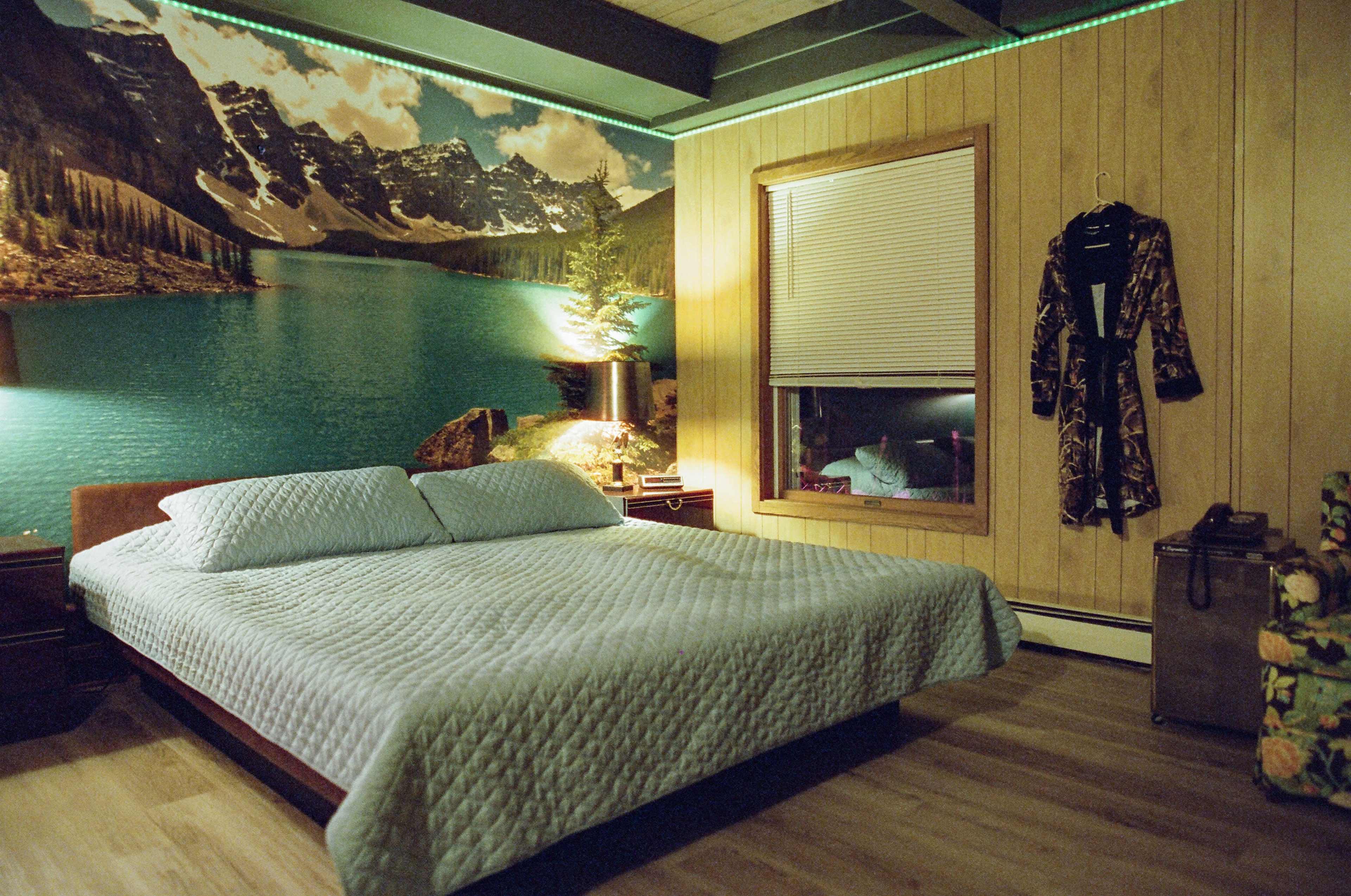 A bedroom with a large bed covered in a light blue quilt, surrounded by wooden paneling and a nature-themed mural of mountains and a lake.