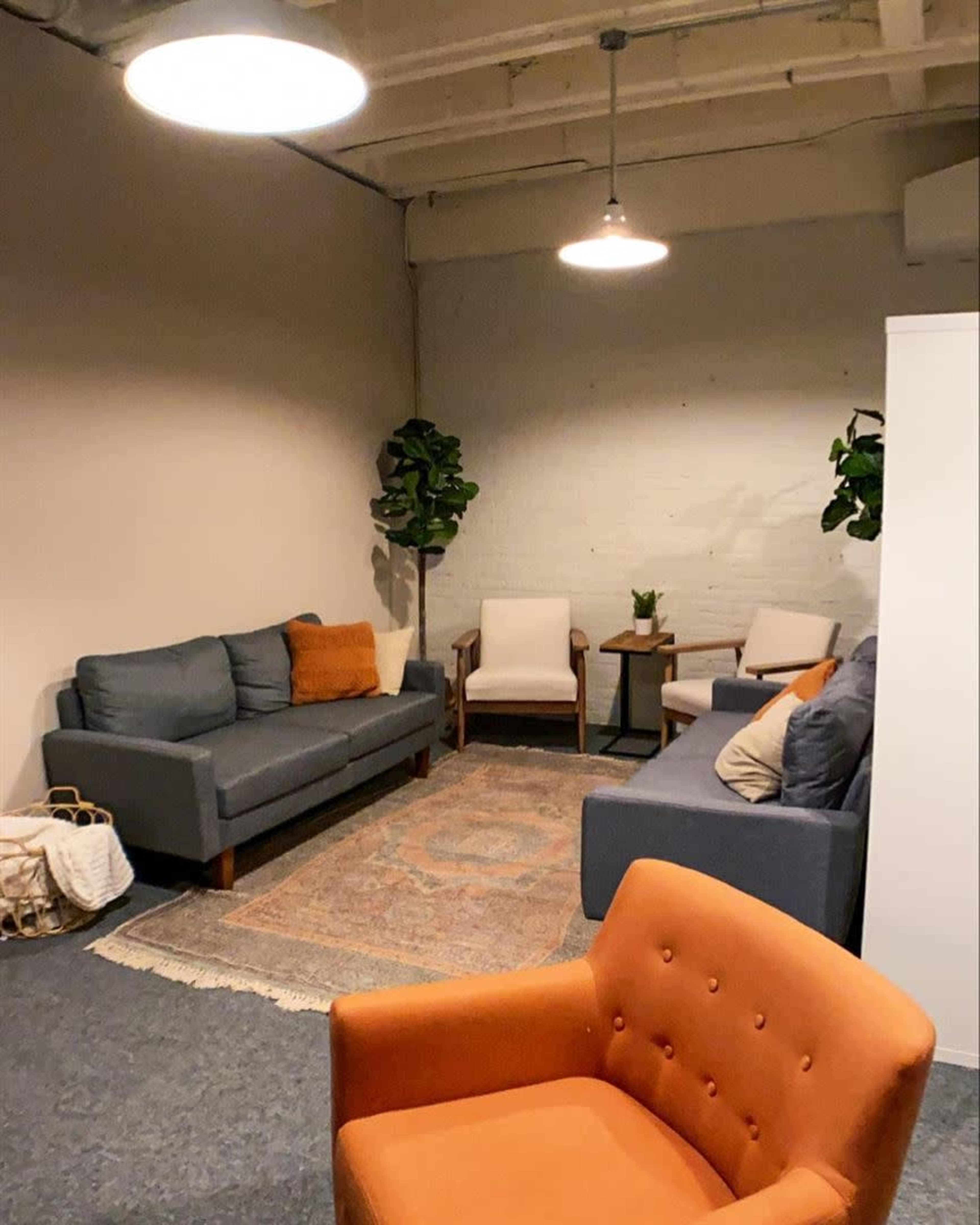 The image shows a cozy lounge area with gray sofas, an orange armchair, a small side table, and green plants in a softly lit room.