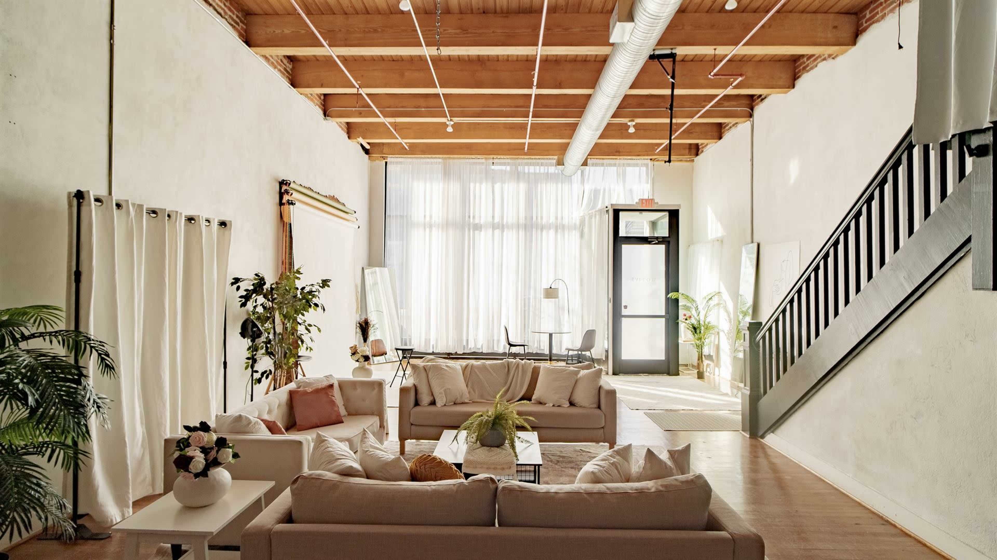 A spacious living area features light-colored sofas arranged around a coffee table, with large windows allowing natural light to fill the room.