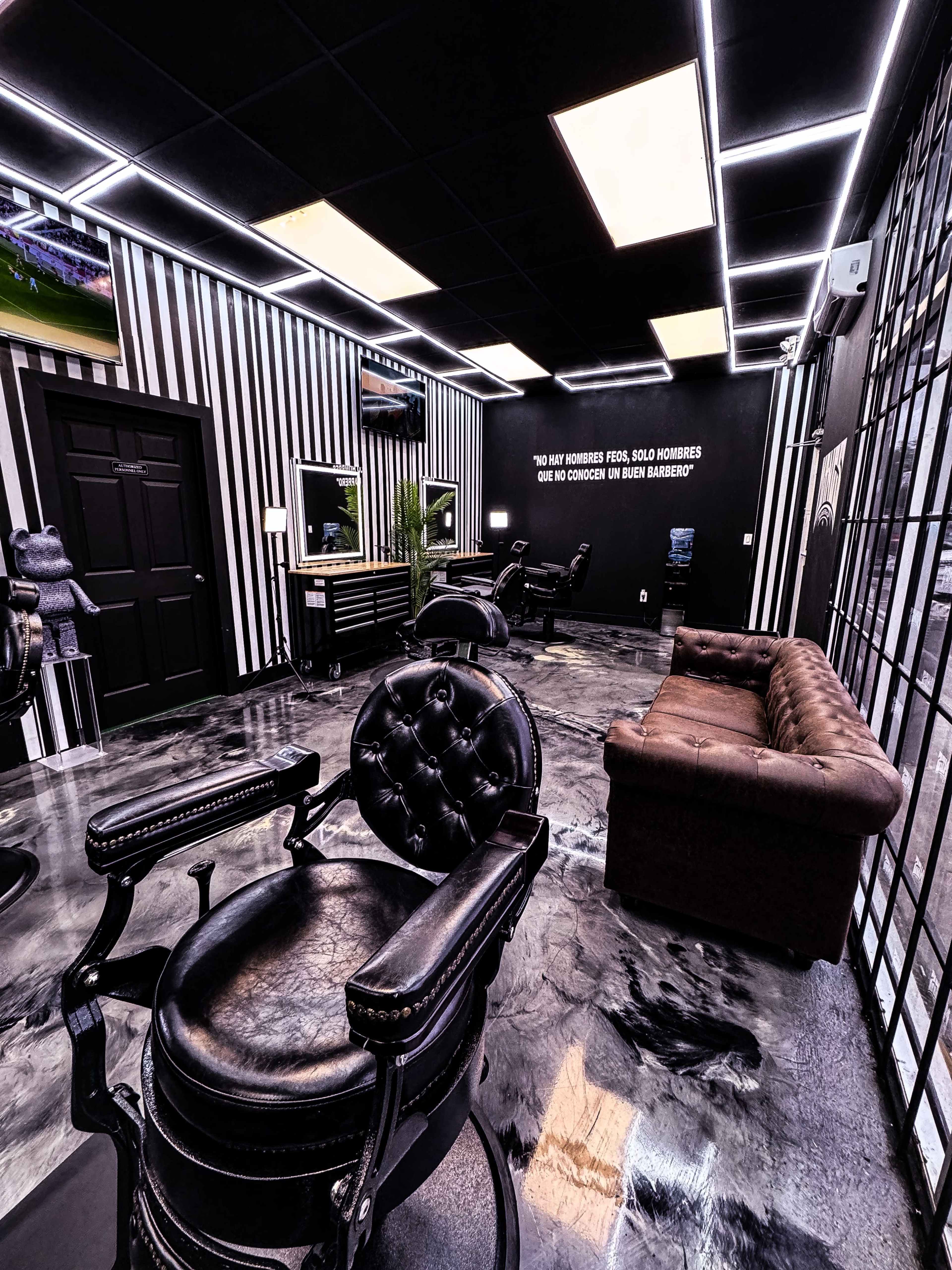 The interior of a modern barbershop features black leather barber chairs, a brown leather couch, and illuminated walls, with a quote displayed on one wall and a television on another.
