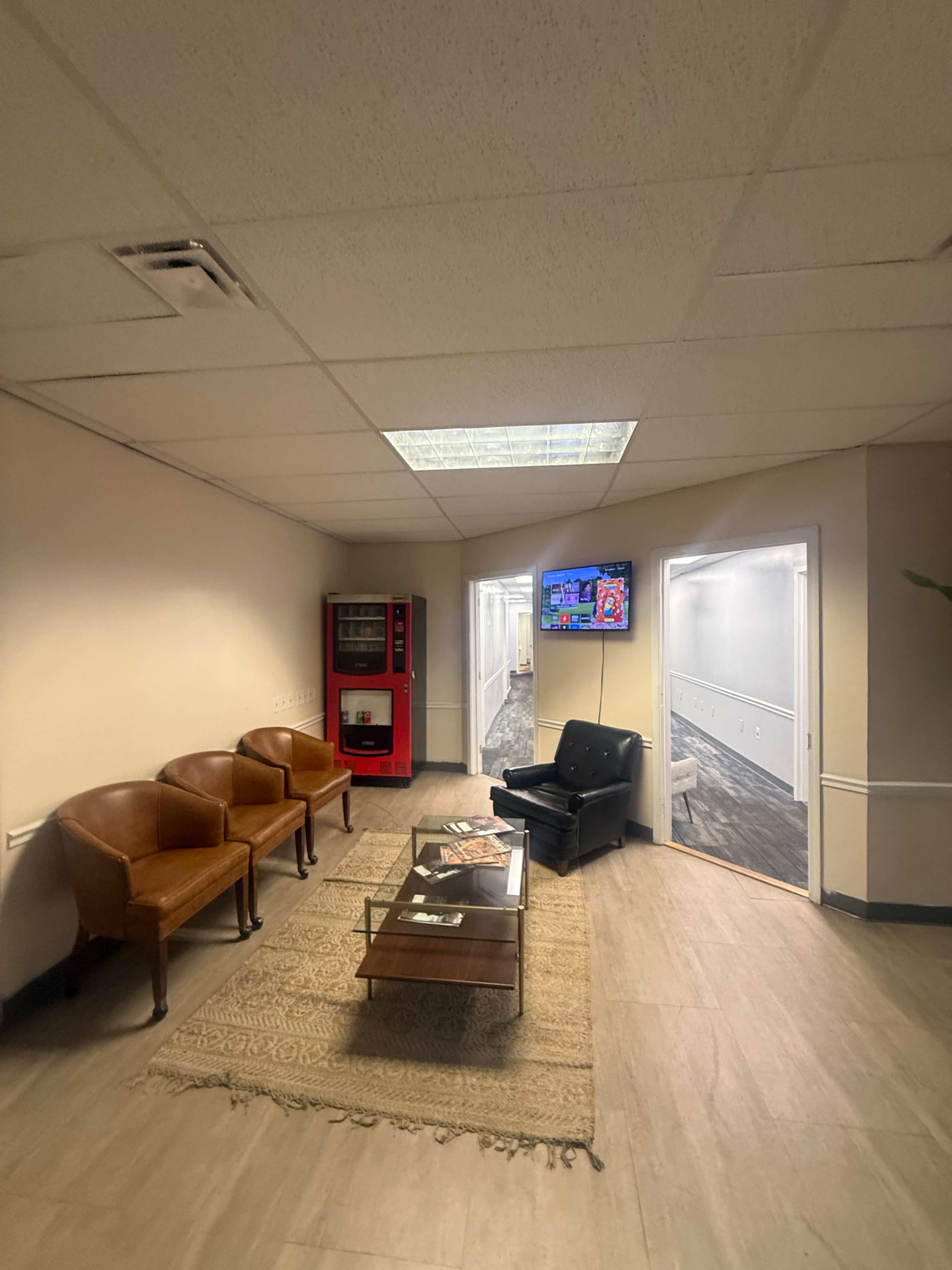 The image shows a hallway with two brown chairs, a black chair, a glass coffee table on a rug, a vending machine, and a television mounted on the wall.