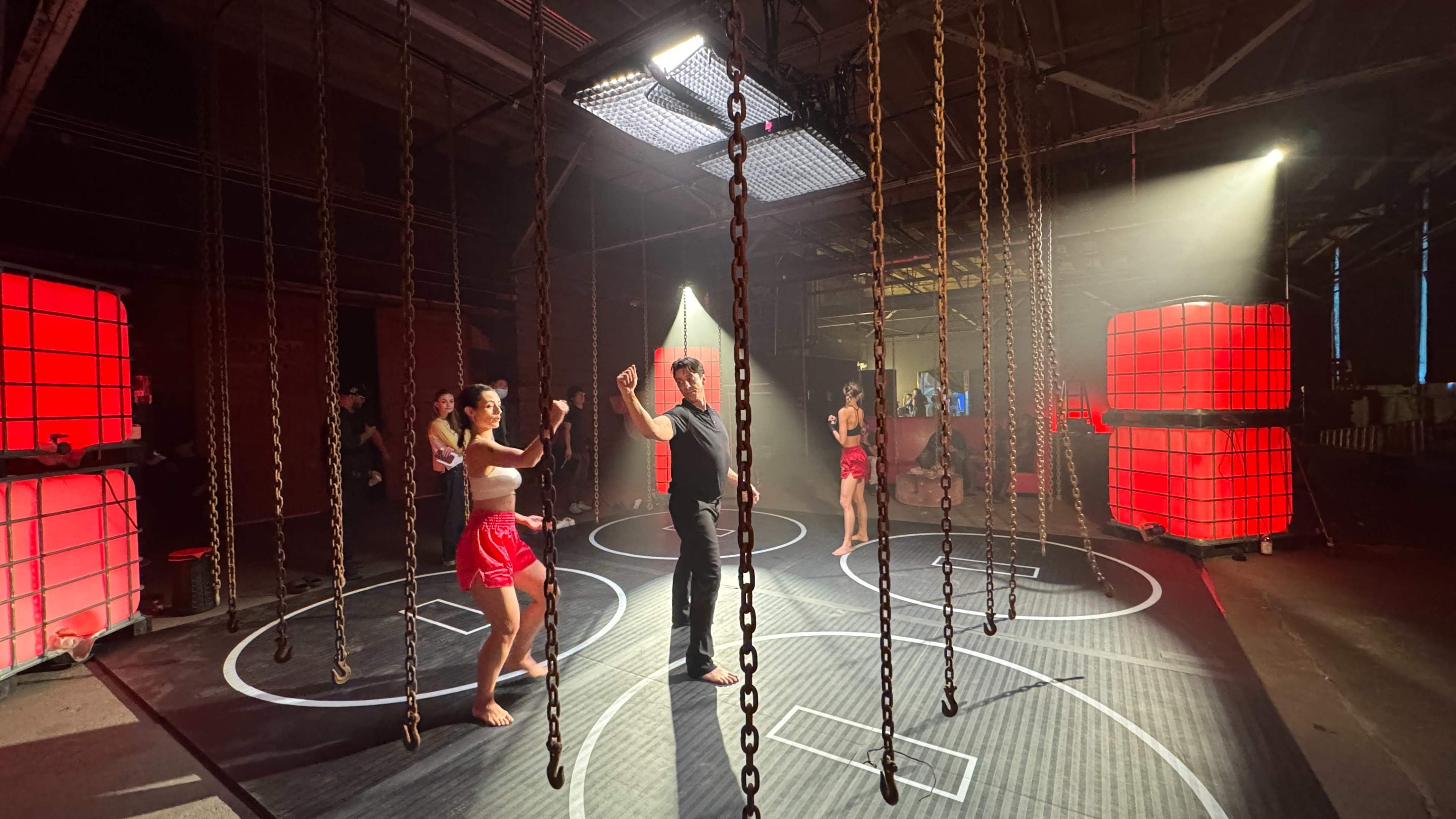 A group of dancers performs in a dimly lit industrial space, surrounded by hanging chains and illuminated by red and white lights.