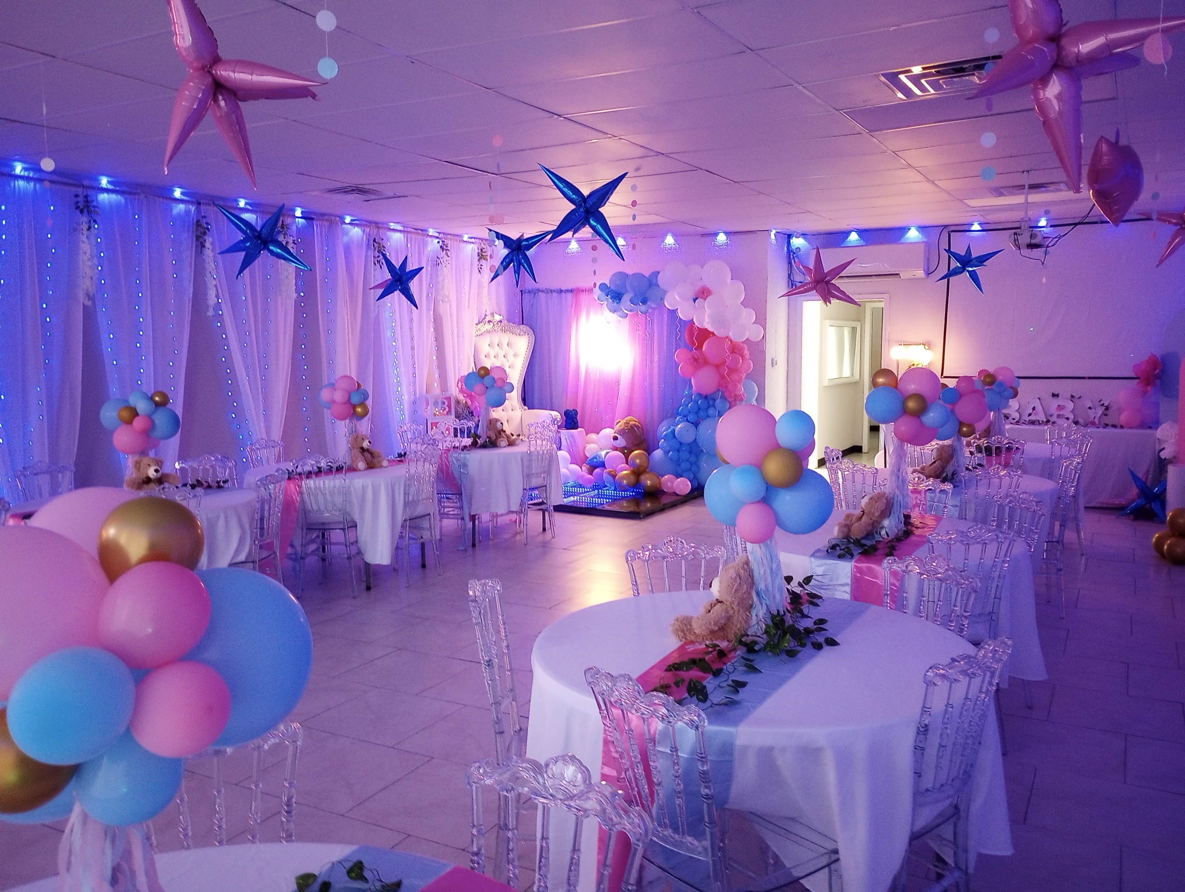 The image shows a brightly decorated party hall with tables set for a celebration, featuring colorful balloons and a backdrop of lights and decorations.
