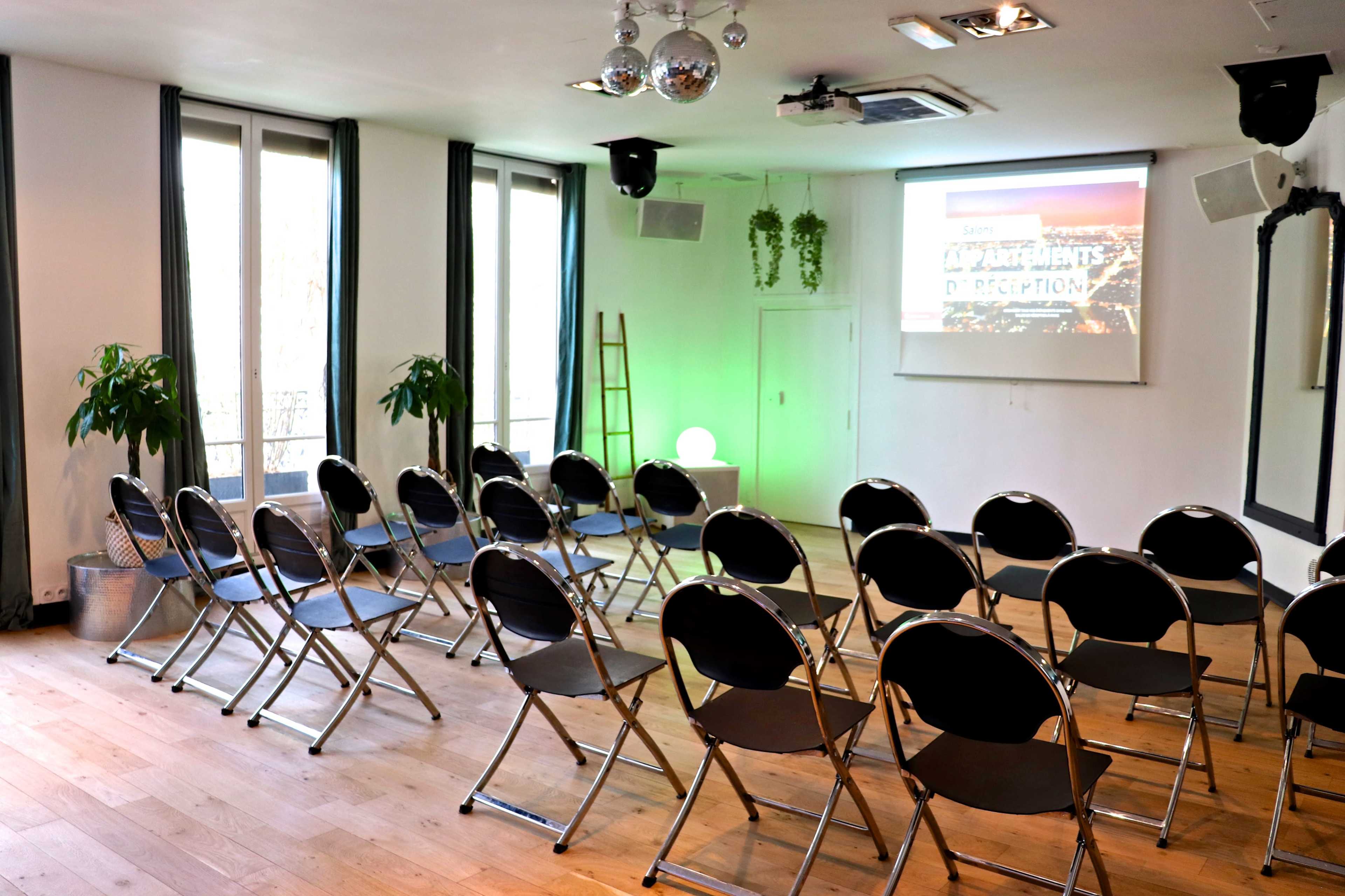 A seminar room is set up with rows of black folding chairs facing a screen, illuminated by a green light, with plants and a ladder in the background.