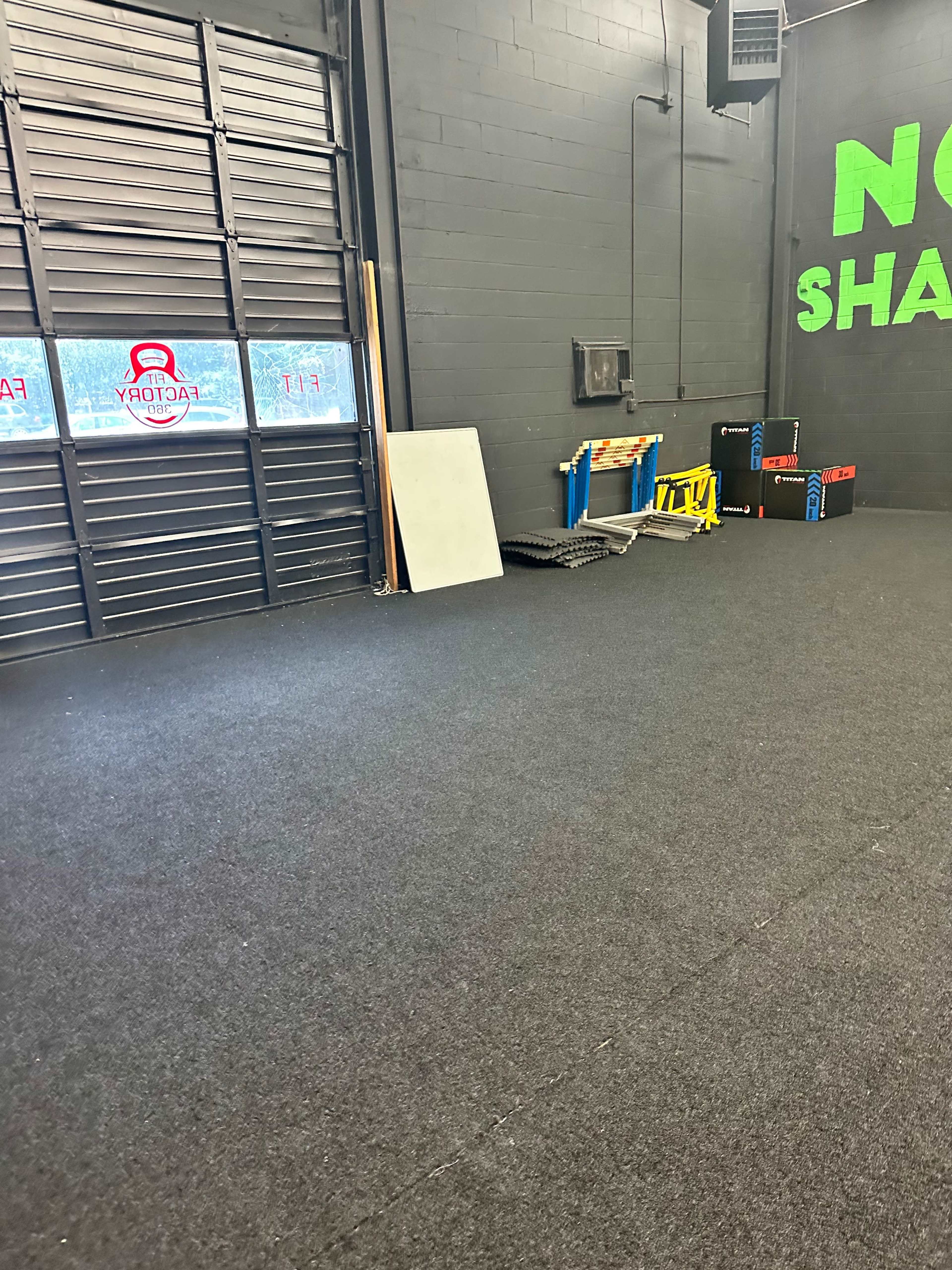 An empty garage-like space with a black carpeted floor, a large closed door with a logo, and several items including a folding table and storage boxes.