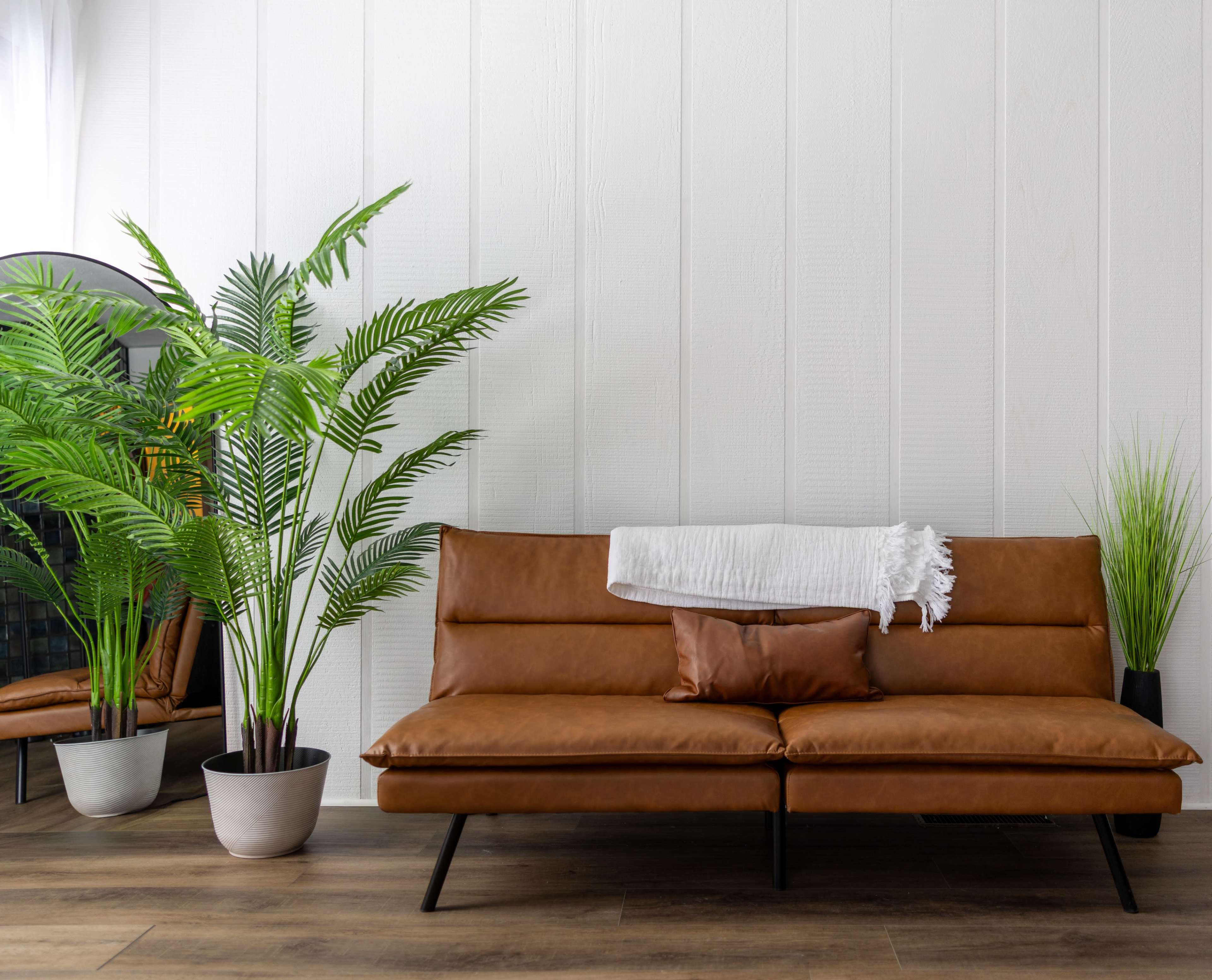 A brown leather sofa with a white throw blanket and a decorative pillow is positioned near potted plants against a white paneled wall.